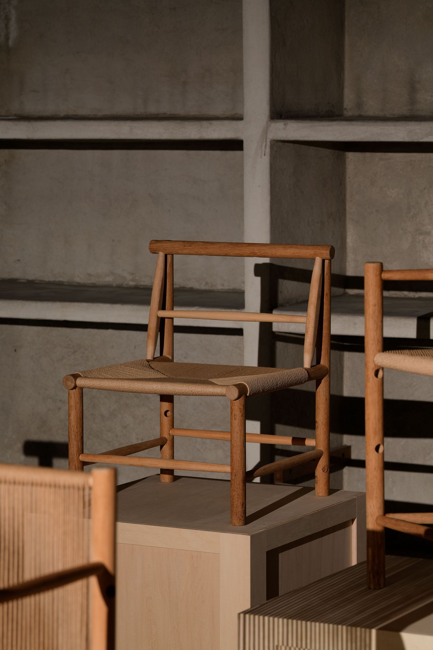 A wooden chair with a woven seat is showcased on a pedestal, with a minimalist background of gray walls and shelves. A wooden chair with a woven seat is showcased on a pedestal, with a minimalist background of gray walls and shelves.