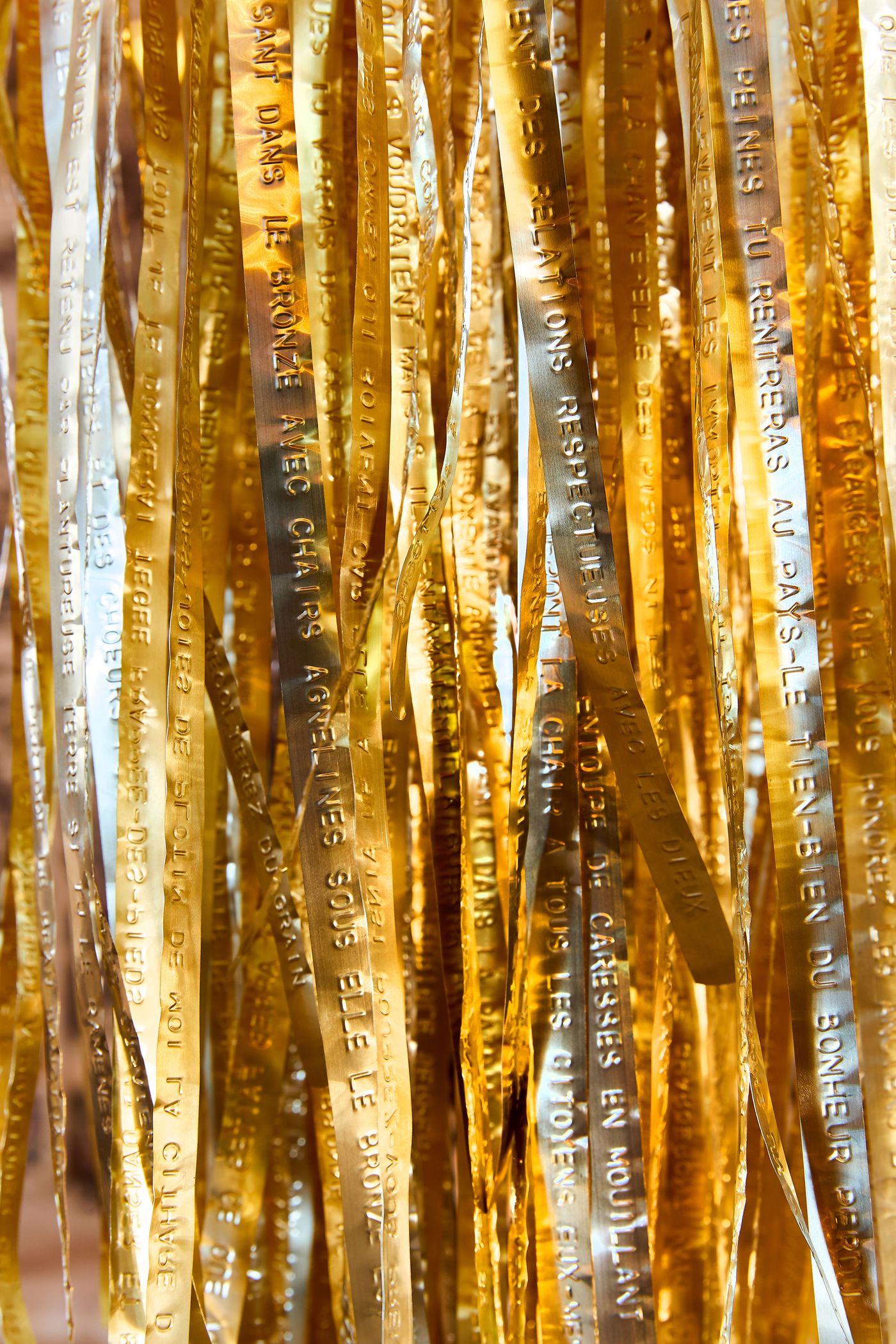 A close-up of a contemporary art installation composed of many vertical, metallic strips in varying shades of silver and gold. The strips are etched with French text, creating a shimmering, layered effect. The light reflects off the polished surfaces, highlighting the interplay between the metallic texture and the engraved phrases.