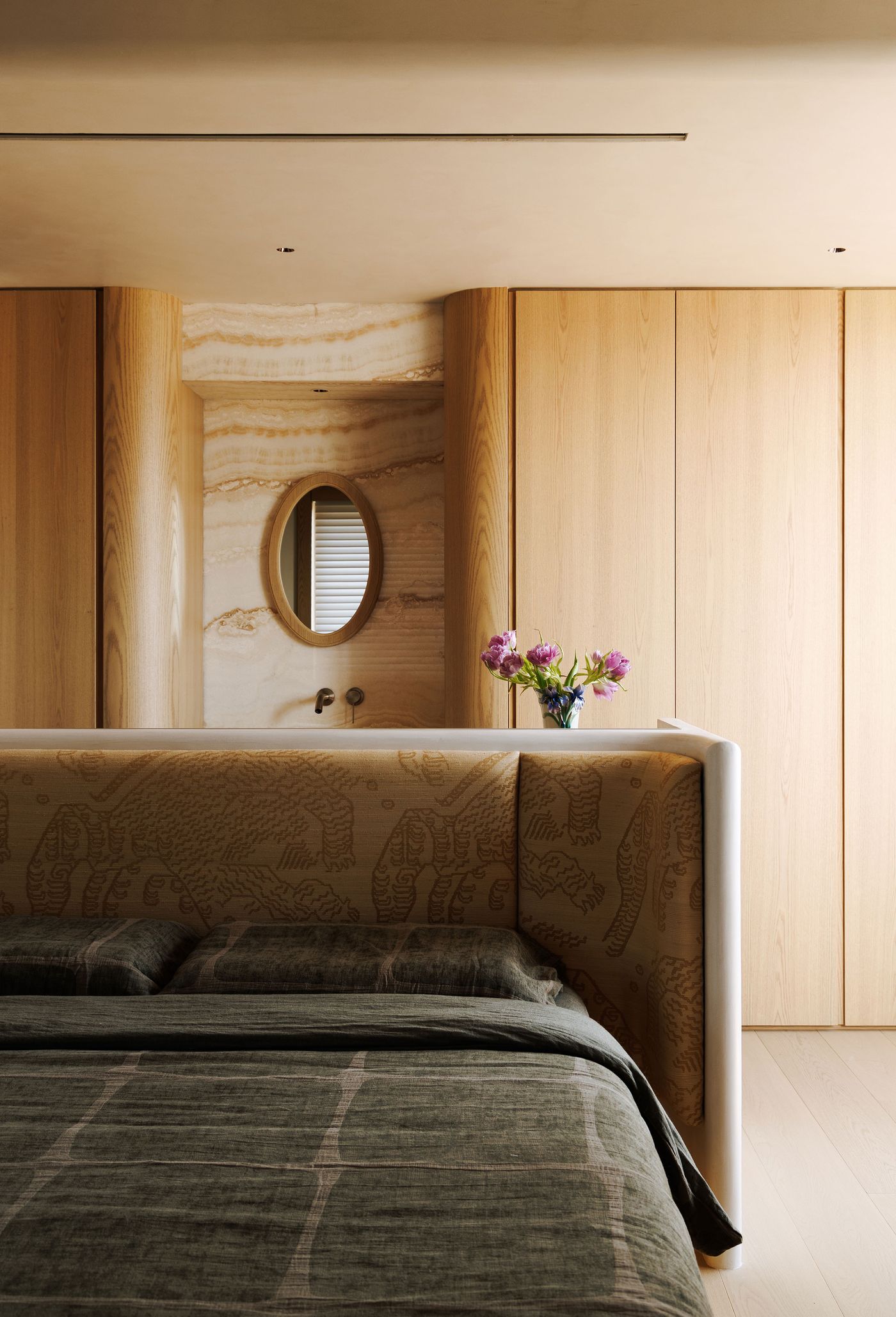 A luxurious and thoughtfully detailed bedroom. The headboard is upholstered in a rich, patterned fabric, creating a focal point. Warm wooden walls and integrated cabinetry surround a vanity area with a distinctive oval mirror set against veined marble, showcasing a blend of natural materials and bespoke craftsmanship for a sophisticated retreat. A luxurious and thoughtfully detailed bedroom. The headboard is upholstered in a rich, patterned fabric, creating a focal point. Warm wooden walls and integrated cabinetry surround a vanity area with a distinctive oval mirror set against veined marble, showcasing a blend of natural materials and bespoke craftsmanship for a sophisticated retreat.