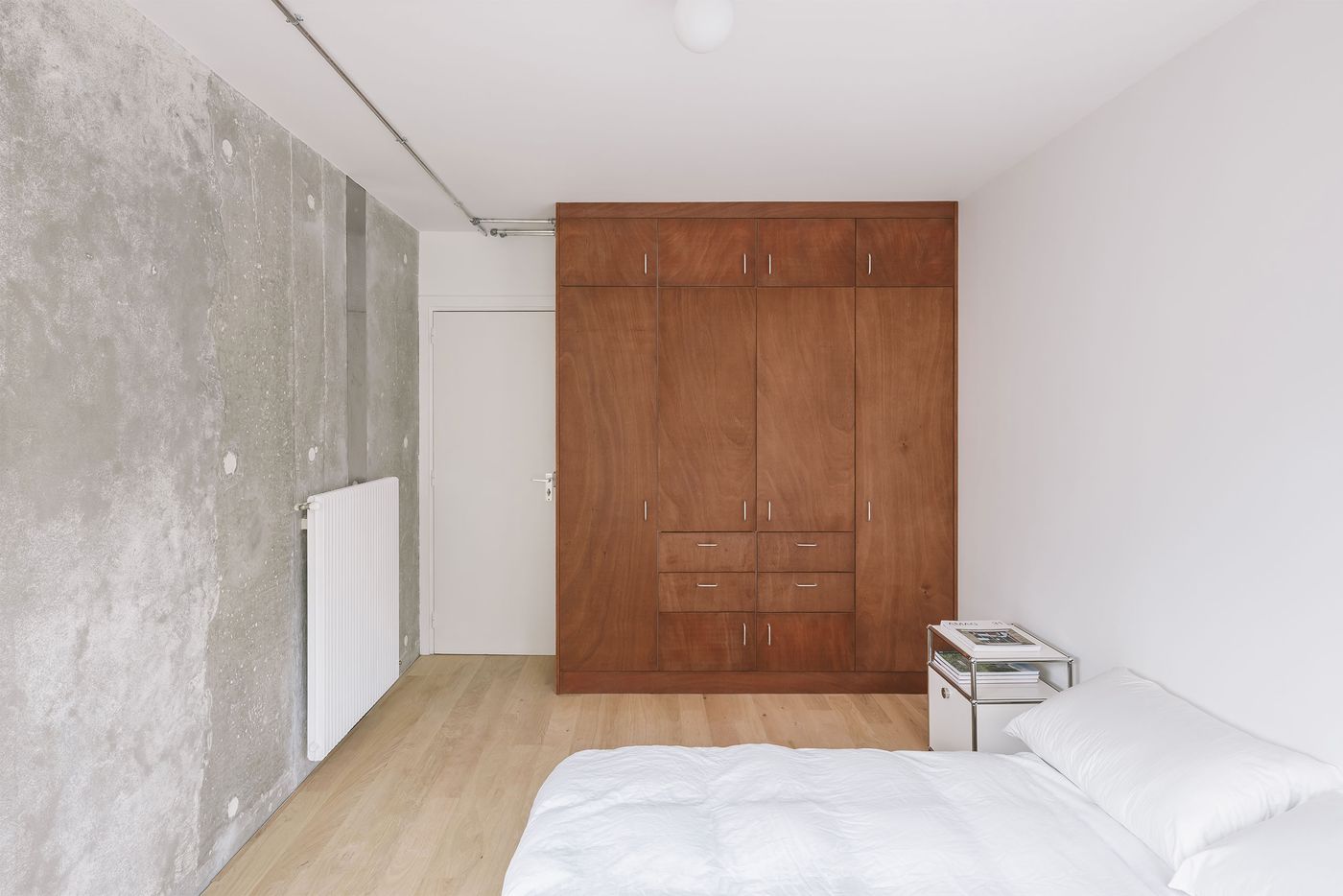 A minimalistic bedroom featuring a wooden wardrobe, concrete wall, white bedding, and a small side table with magazines.
