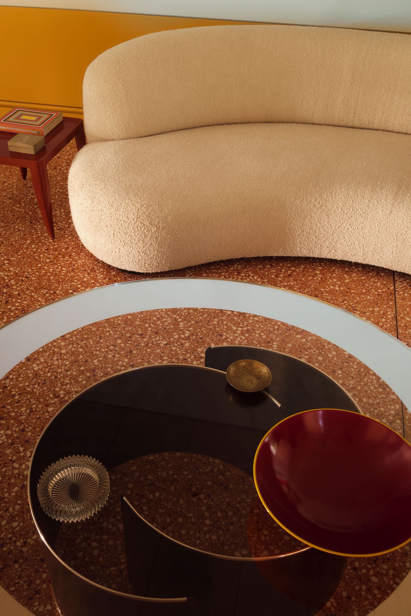 A cozy, curved beige sofa beside a sleek coffee table adorned with elegant bowls, set on textured flooring and accented by a vibrant wall.
