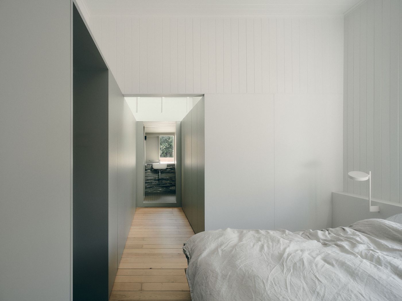A minimalist bedroom features a wooden floor, soft bedding, and a sleek lamp, leading to a bright hallway with a bathroom at the end. A minimalist bedroom features a wooden floor, soft bedding, and a sleek lamp, leading to a bright hallway with a bathroom at the end.