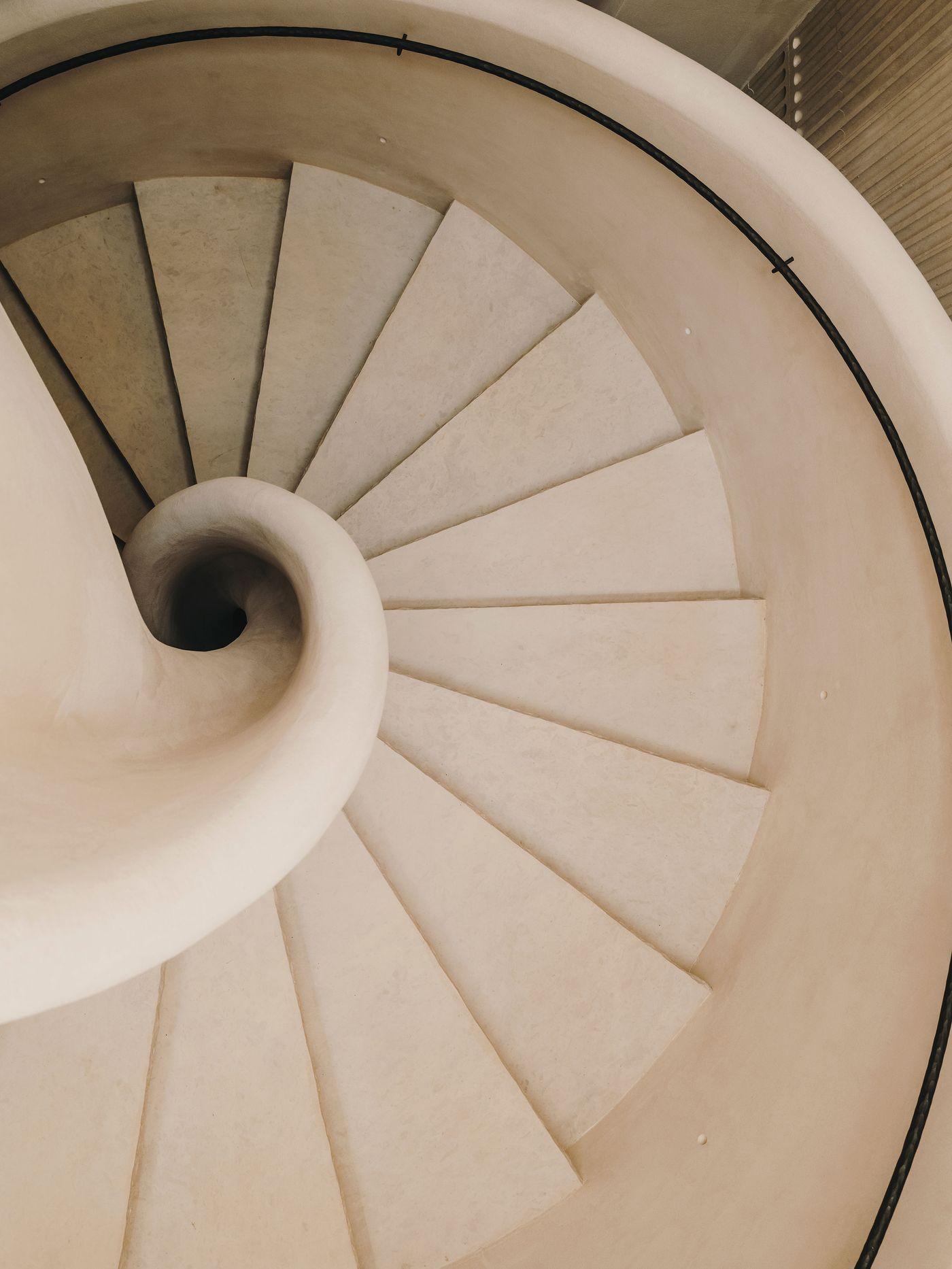 Aerial view of a spiral staircase with smooth, beige steps and a minimalist black handrail curving elegantly around.