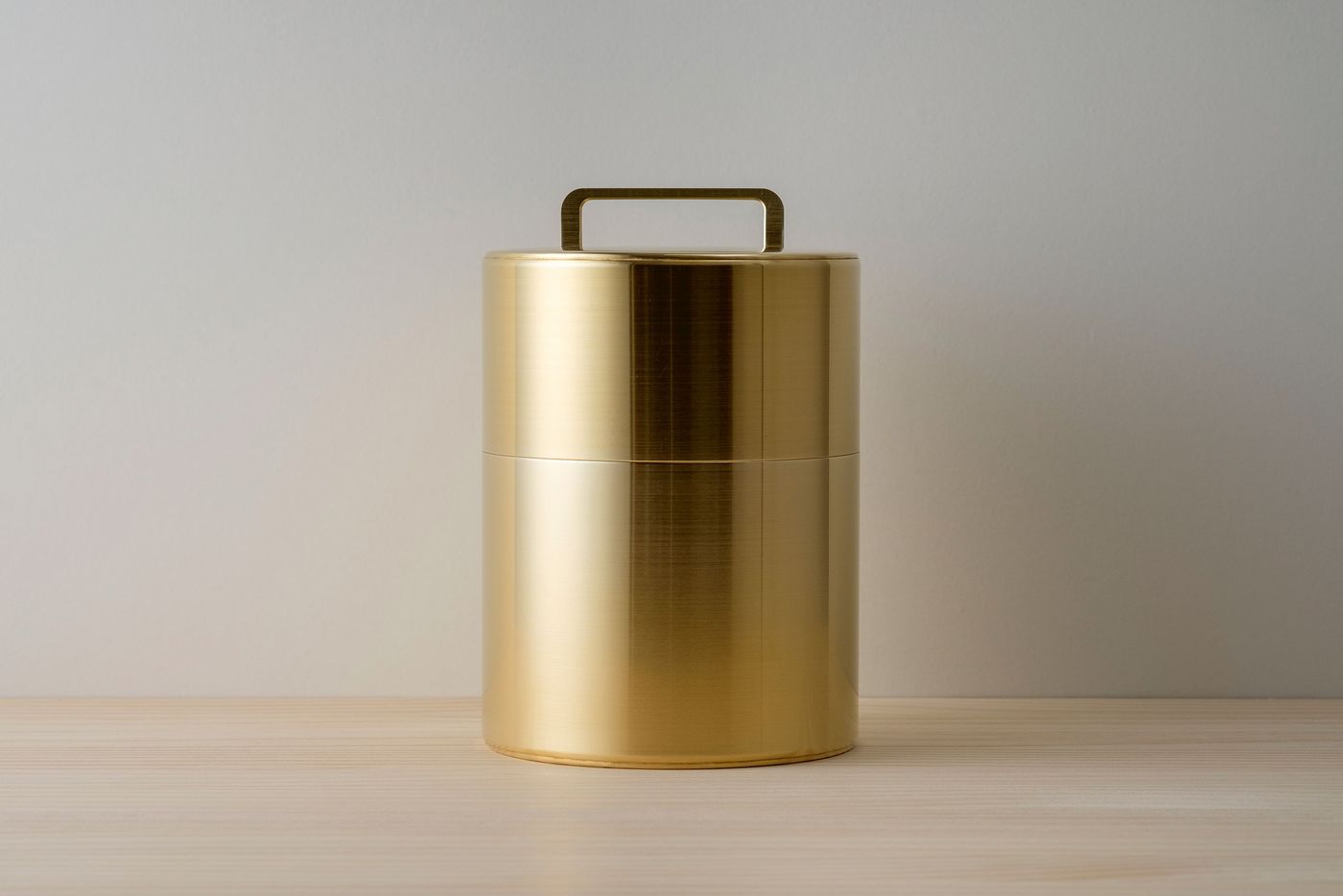 A sleek, gold cylindrical canister with a handle, resting on a light wooden surface against a soft gray background. A sleek, gold cylindrical canister with a handle, resting on a light wooden surface against a soft gray background.