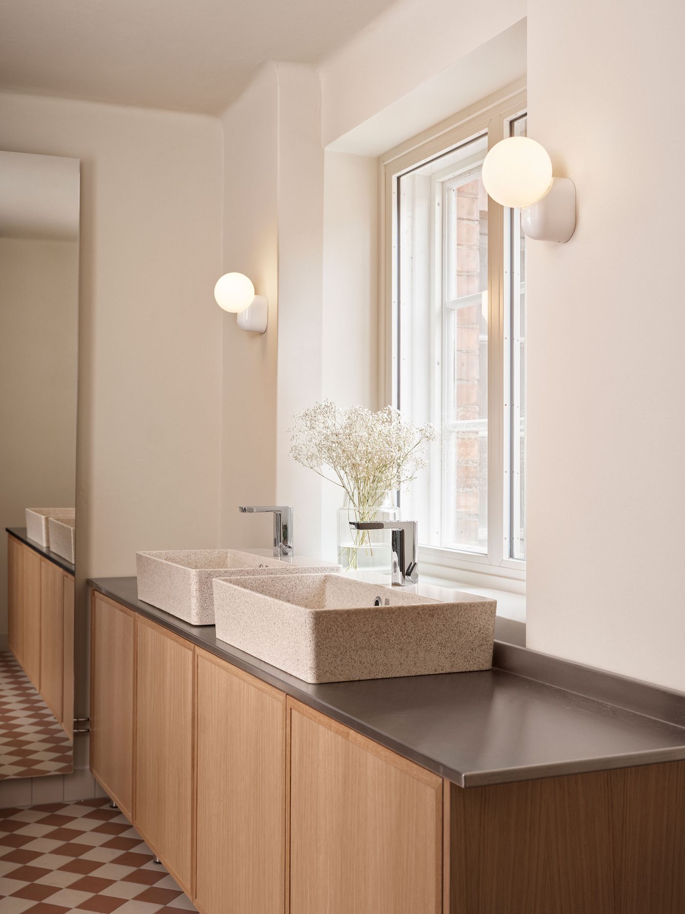 A modern bathroom features light wood cabinetry, stone-like rectangular sinks, and sleek faucets, all illuminated by spherical wall-mounted lights. The distinctive red and white checkerboard floor adds a playful touch to the clean, contemporary design.