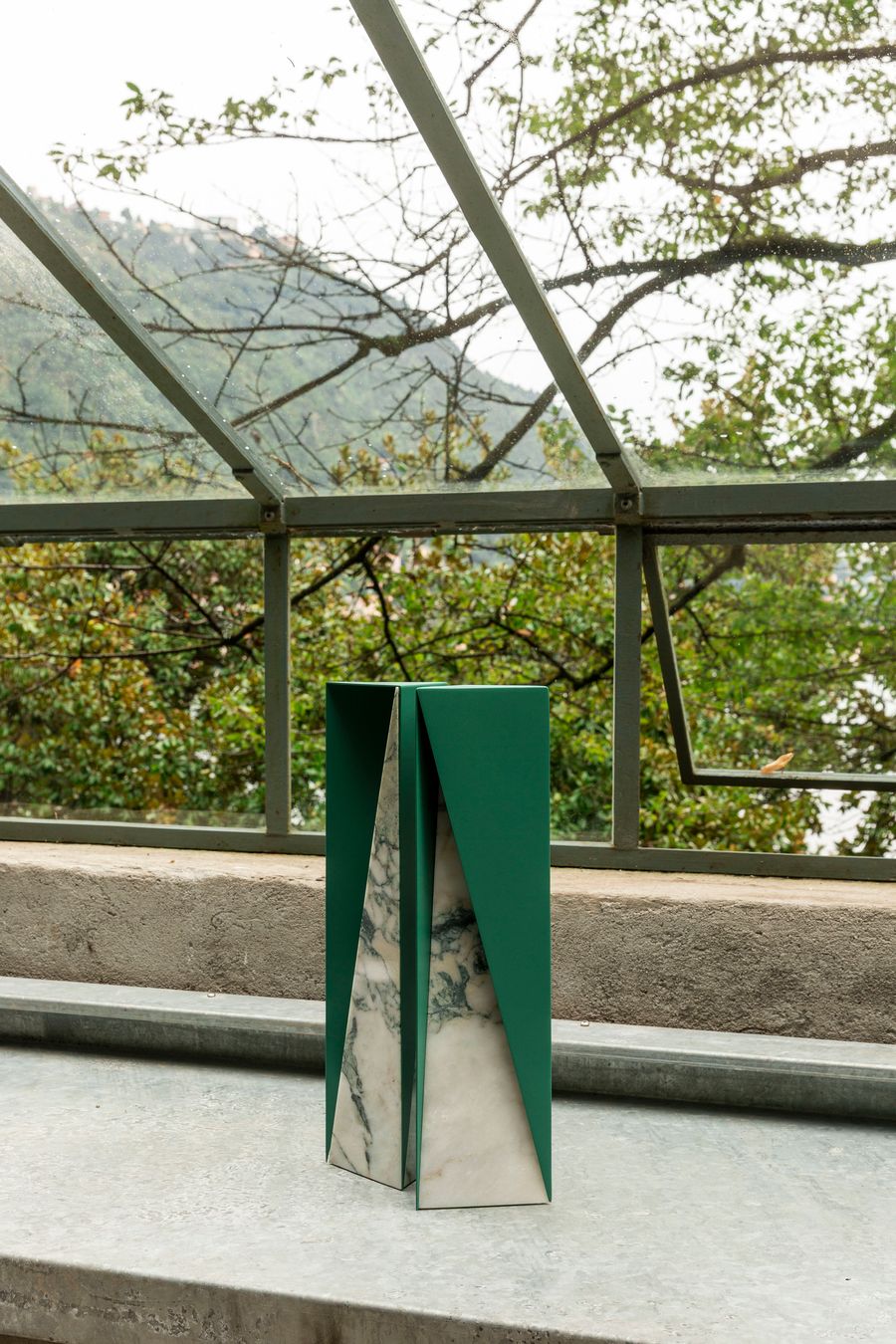 A striking geometric pedestal combines deep green metal panels with a striking marbled insert, forming a sculptural form. It stands on a concrete ledge, with a blurred background of trees and mountains seen through the glass panes of a conservatory-like structure. The bold colors and sharp lines of the object contrast with the soft, organic textures of the natural environment, creating a modern and artistic statement.