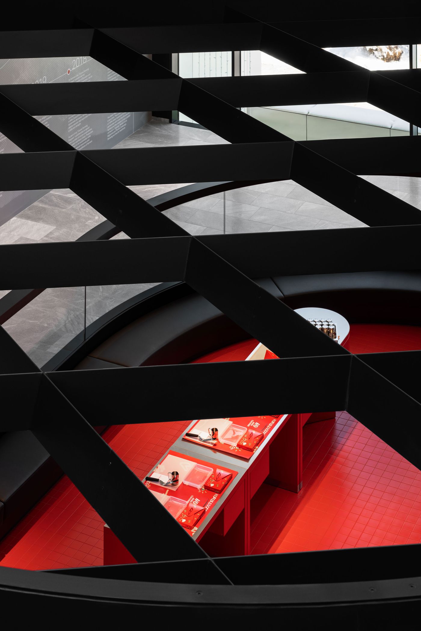 Geometric black ceiling structure creates dramatic shadows above a curved exhibition space, with glimpses of red presentation areas below. The interplay between angular overhead elements and flowing ground-level forms demonstrates sophisticated spatial layering, where architecture becomes both frame and participant in the visitor experience. Geometric black ceiling structure creates dramatic shadows above a curved exhibition space, with glimpses of red presentation areas below. The interplay between angular overhead elements and flowing ground-level forms demonstrates sophisticated spatial layering, where architecture becomes both frame and participant in the visitor experience.