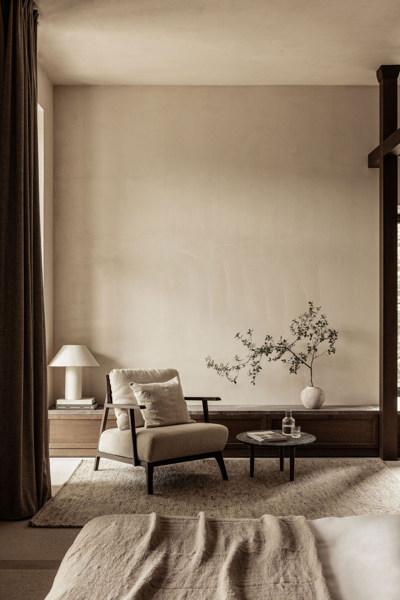 A tranquil and minimalist living area, adjacent to a bed, features a comfortable, low-slung armchair with a plush cushion. Soft, natural light illuminates the textured, neutral-toned walls and a subtle dark timber base. A delicate branch in a ceramic vase and a contemporary table lamp complete the serene, understated aesthetic. A tranquil and minimalist living area, adjacent to a bed, features a comfortable, low-slung armchair with a plush cushion. Soft, natural light illuminates the textured, neutral-toned walls and a subtle dark timber base. A delicate branch in a ceramic vase and a contemporary table lamp complete the serene, understated aesthetic.