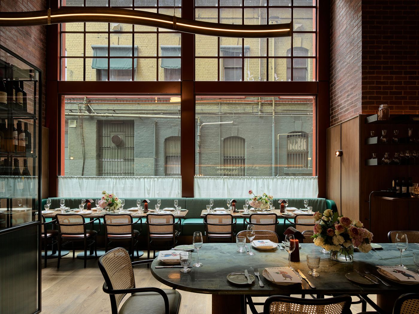 An elegantly set restaurant with a large window view, featuring tables adorned with flowers and glassware against a backdrop of brick and greenery.