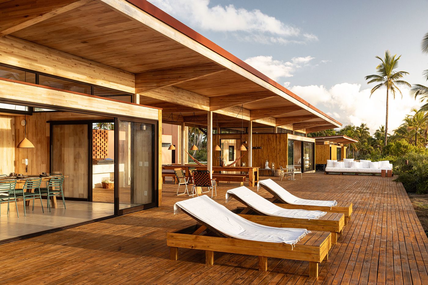 A wide view of a low-slunf, timber-clad complex stretching along a wooden deck, featuring open dining areas and chaise lounges, all under a vast overhanging roof. The seamless integration with the tropical environment and casual, resort-like amenities reflect a sophisticated coastal design. A wide view of a low-slunf, timber-clad complex stretching along a wooden deck, featuring open dining areas and chaise lounges, all under a vast overhanging roof. The seamless integration with the tropical environment and casual, resort-like amenities reflect a sophisticated coastal design.