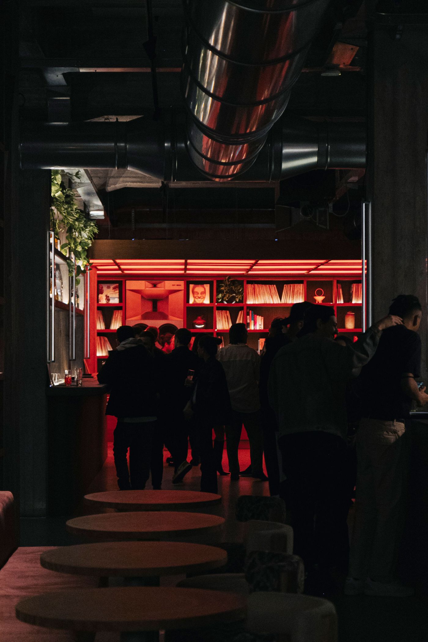 A lively, dimly lit bar scene with a vibrant red bookshelf backdrop, patrons mingling, and round tables in the foreground. A lively, dimly lit bar scene with a vibrant red bookshelf backdrop, patrons mingling, and round tables in the foreground.