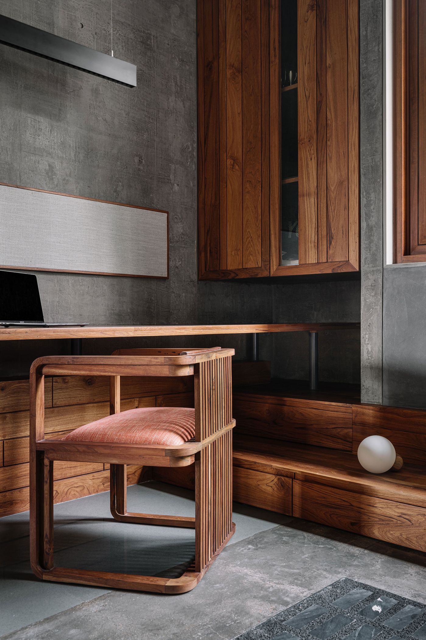 A refined home office nook, seamlessly integrated into a larger space, defined by a bespoke wooden desk and a slatted wooden chair with a subtly contrasting upholstered seat. Rich wooden cabinetry lines the wall, while a long, white rectangular art piece hangs above the desk. The raw concrete wall provides an industrial backdrop.