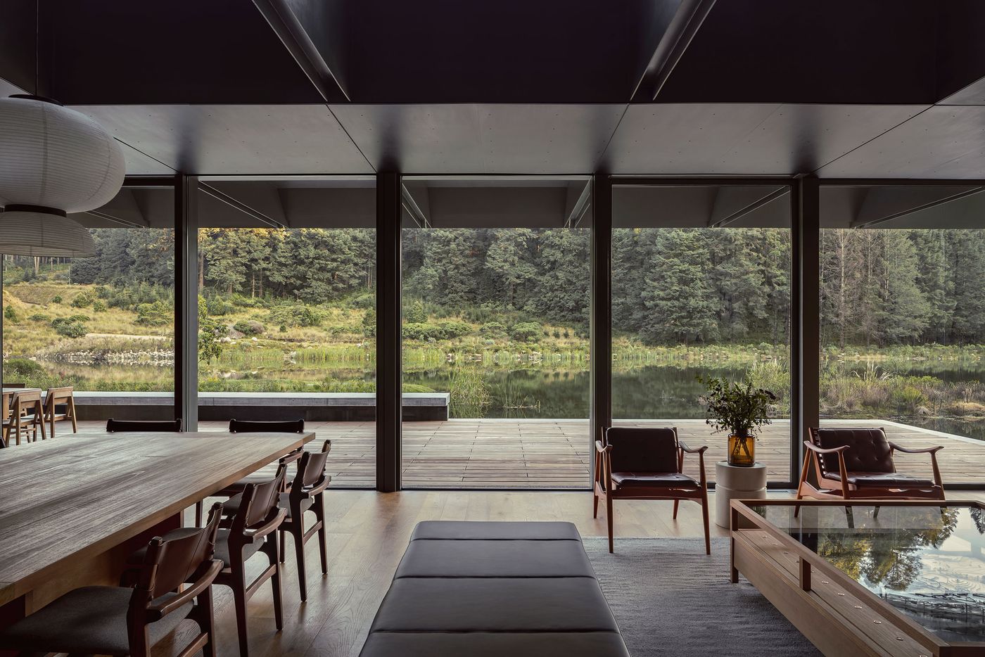 Modern, minimalist interior with large windows overlooking a serene lake, framed by lush greenery and contemporary furnishings.