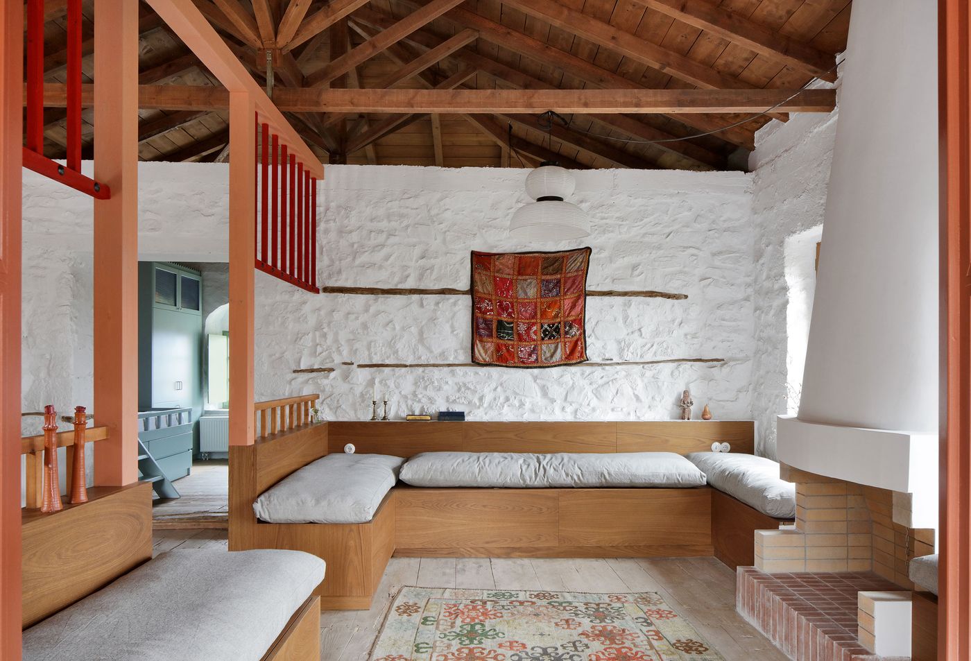 Built-in oak sleeping platforms create intimate alcoves beneath the coral-red mezzanine structure, where traditional patchwork textiles add warmth to the minimalist intervention. The design celebrates both solitude and community through thoughtful spatial subdivision within the historic stone envelope.