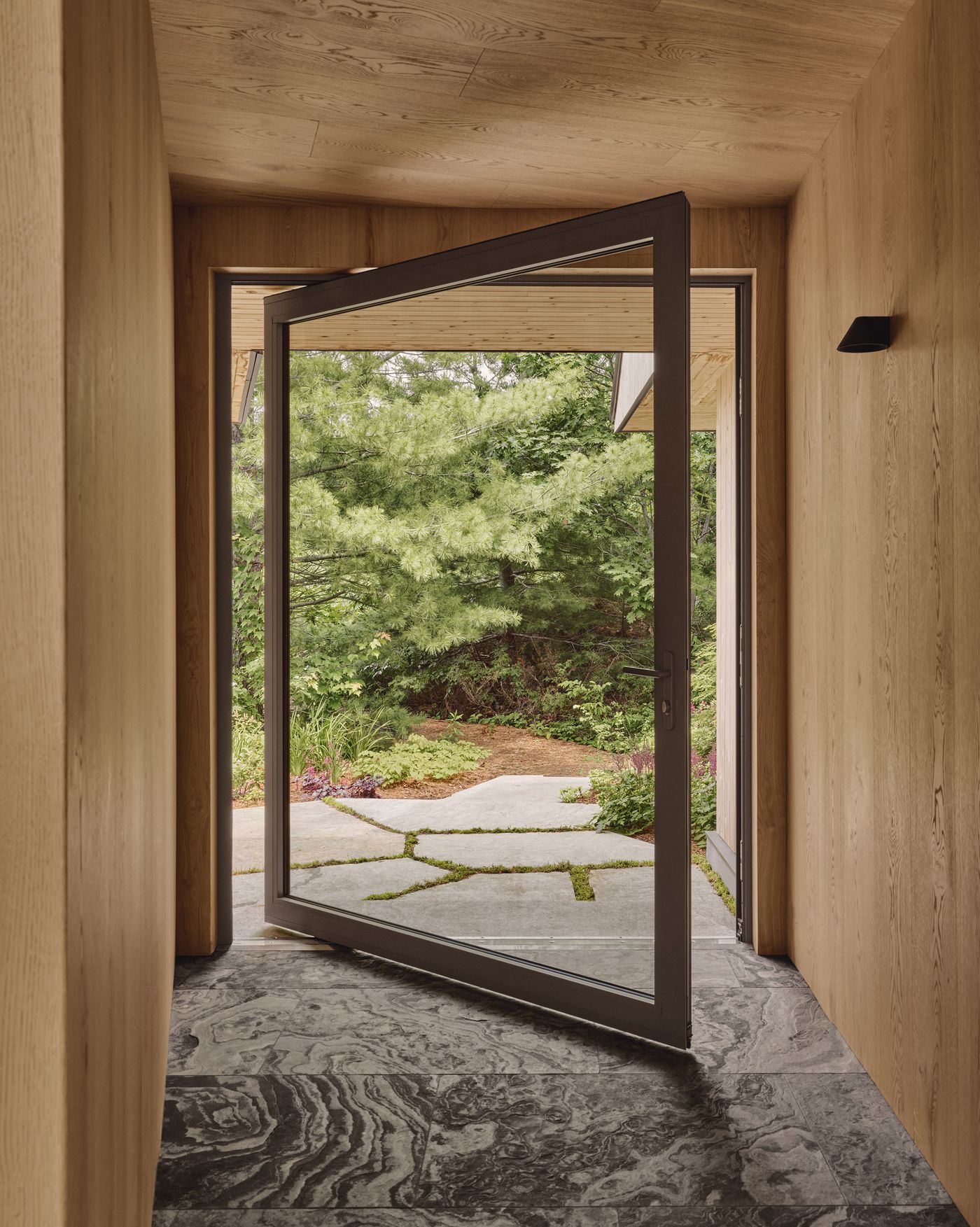 A modern entrance with a large open door, revealing lush greenery and a stone pathway framed by wooden walls and flooring. A modern entrance with a large open door, revealing lush greenery and a stone pathway framed by wooden walls and flooring.
