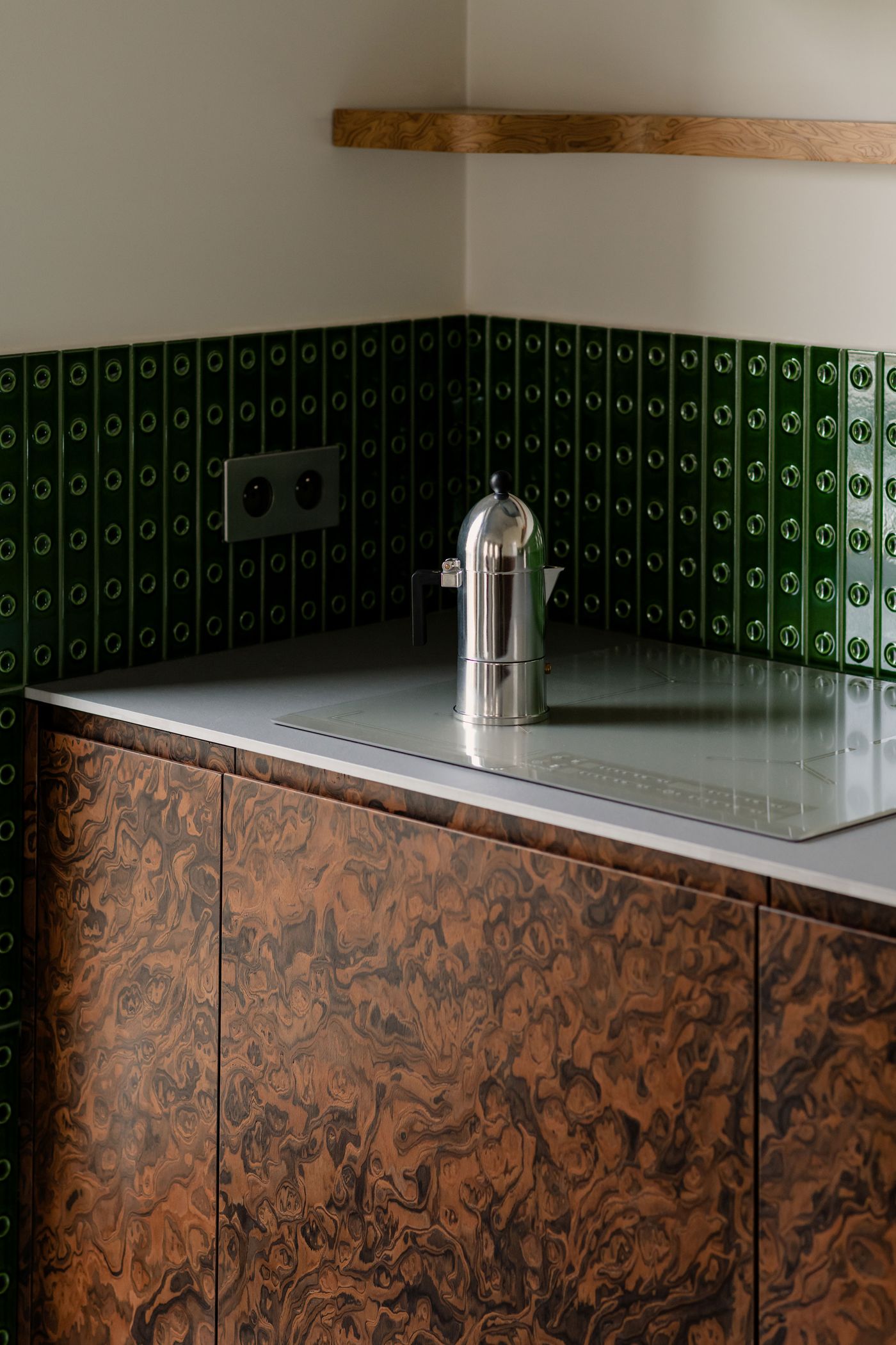 A detail of the kitchen counter reveals the interplay between the reflective metallic moka pot, bottle-green grid tiles, and swirling wood cabinetry—an ode to tactile contrasts and material depth.