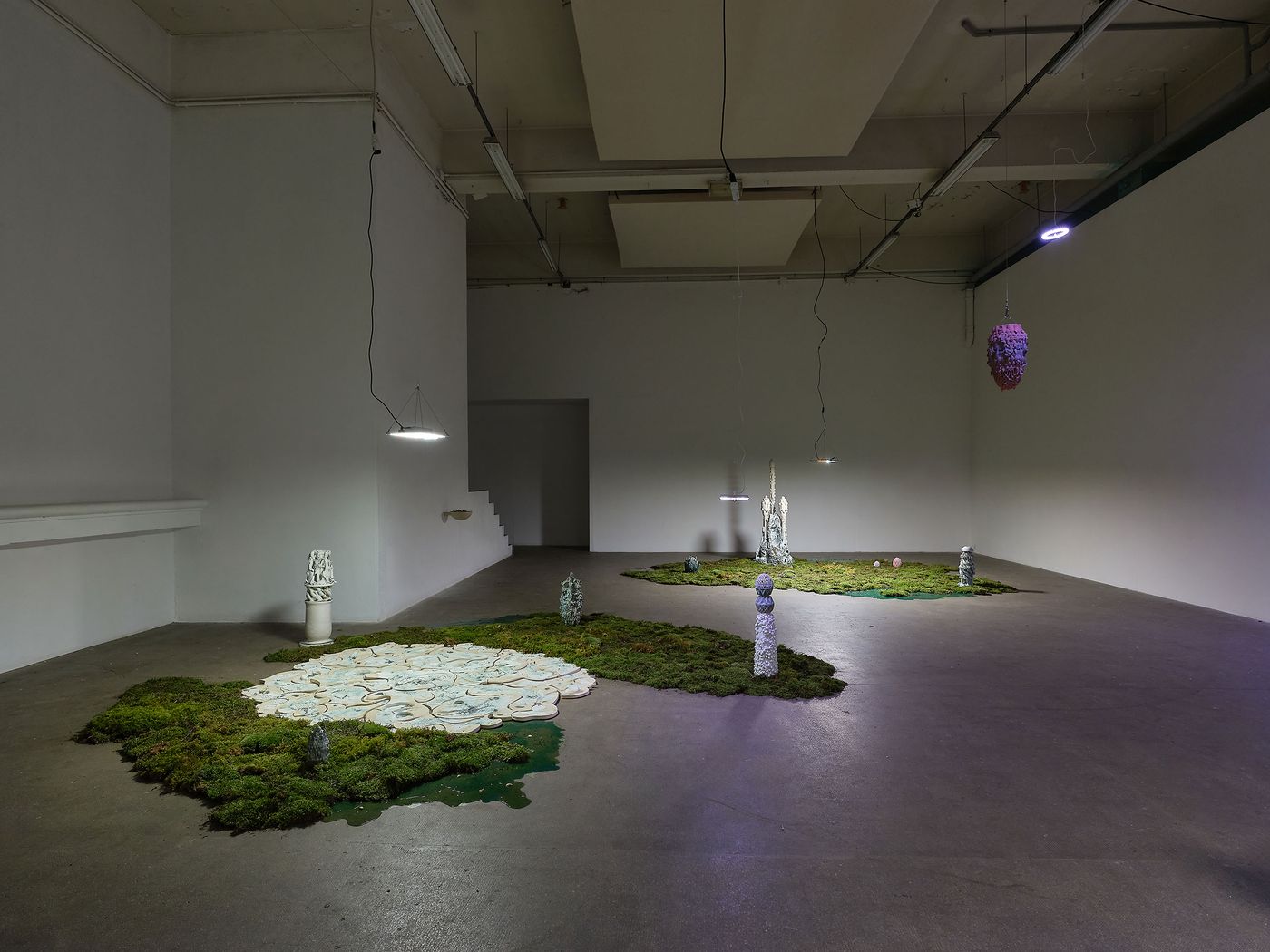 An art installation featuring moss-covered platforms, sculptural forms, and hanging lights in a minimalist gallery space. An art installation featuring moss-covered platforms, sculptural forms, and hanging lights in a minimalist gallery space.