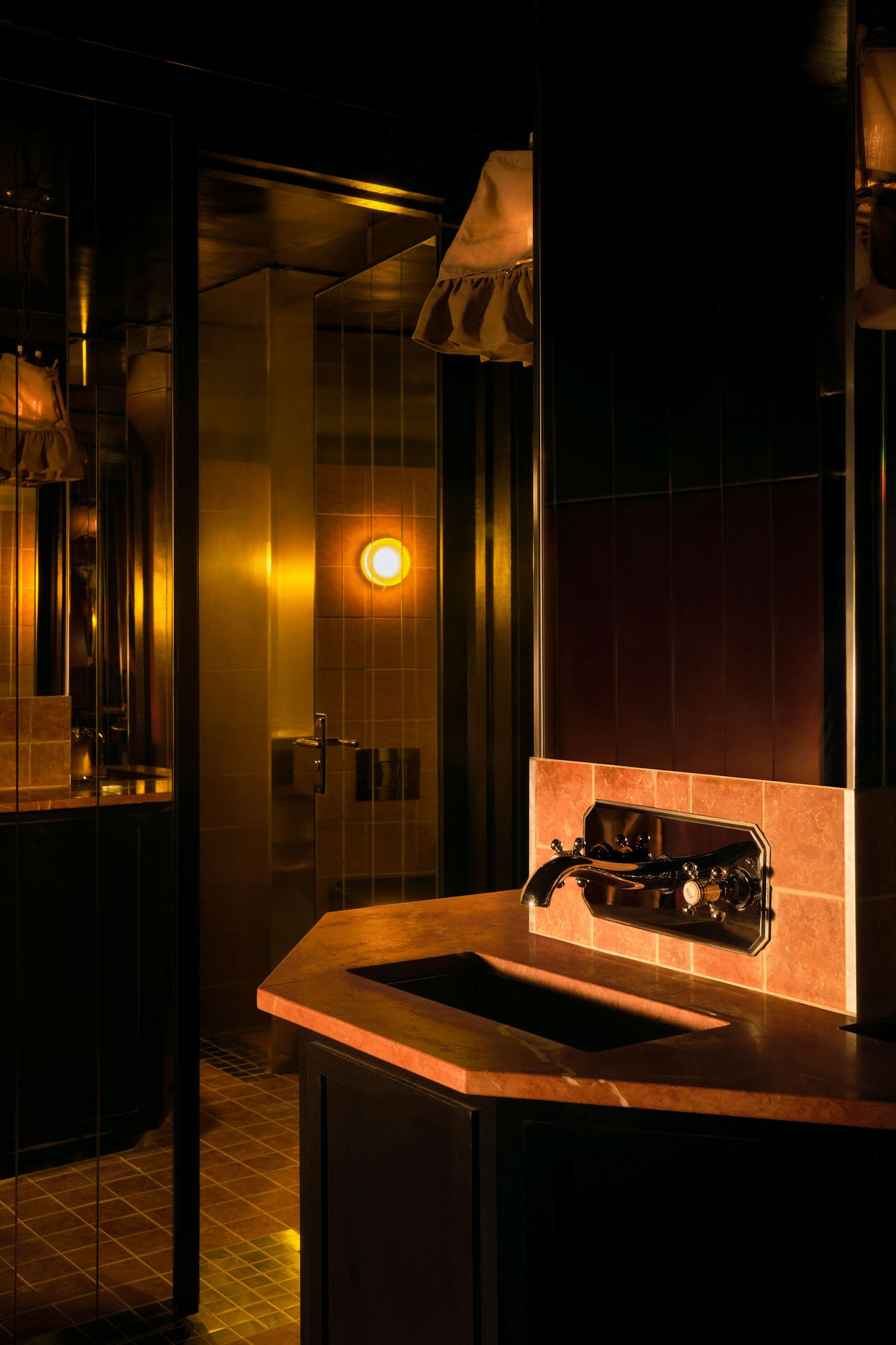 A close-up of a luxurious bathroom counter in The Roses of Elagabalus, featuring a deep red marble countertop with an integrated dark basin and an ornate, vintage-style wall-mounted faucet in dark metal. Mirrored surfaces and a warm amber wall light in the background create a rich, moody, and sophisticated ambiance.