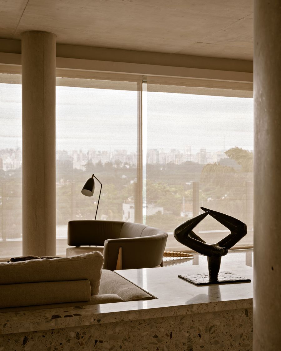 A modern living room features a sleek chair, abstract sculpture, and city view through sheer curtains, with warm, neutral tones. A modern living room features a sleek chair, abstract sculpture, and city view through sheer curtains, with warm, neutral tones.