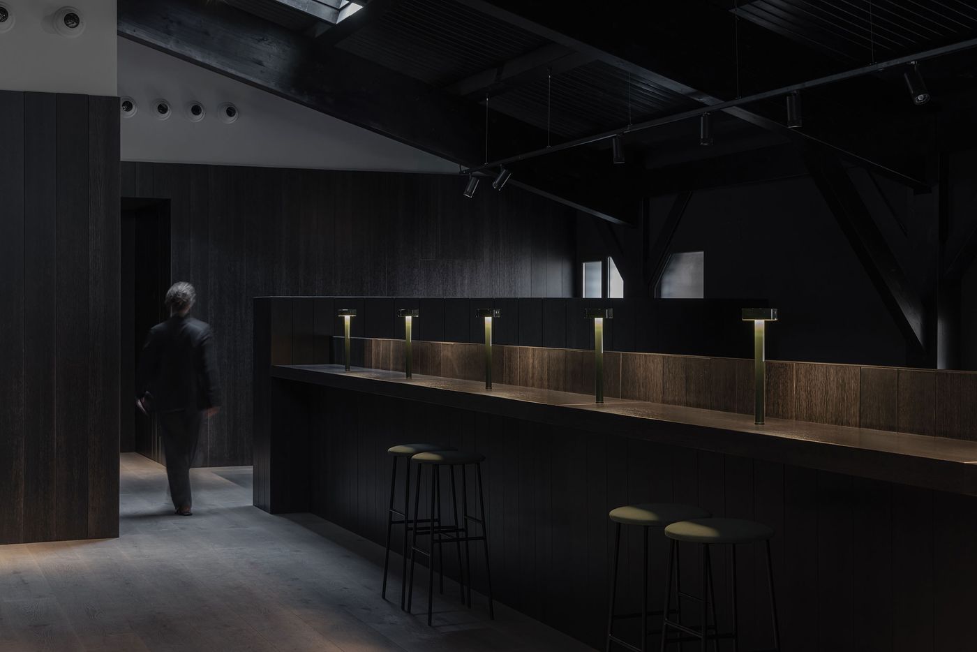 A dimly lit modern interior features a sleek bar with stylish lamps and two stools, as a person walks past a doorway. A dimly lit modern interior features a sleek bar with stylish lamps and two stools, as a person walks past a doorway.