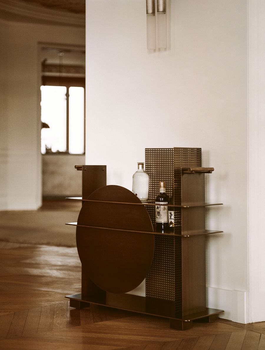A distinctive, sculptural bar cart in dark wood and perforated metal features a striking circular element and cantilevered shelves. Adorned with bottles and a white vase, this piece exemplifies a blend of brutalist and art-deco influences, serving as both furniture and a work of art. A distinctive, sculptural bar cart in dark wood and perforated metal features a striking circular element and cantilevered shelves. Adorned with bottles and a white vase, this piece exemplifies a blend of brutalist and art-deco influences, serving as both furniture and a work of art.