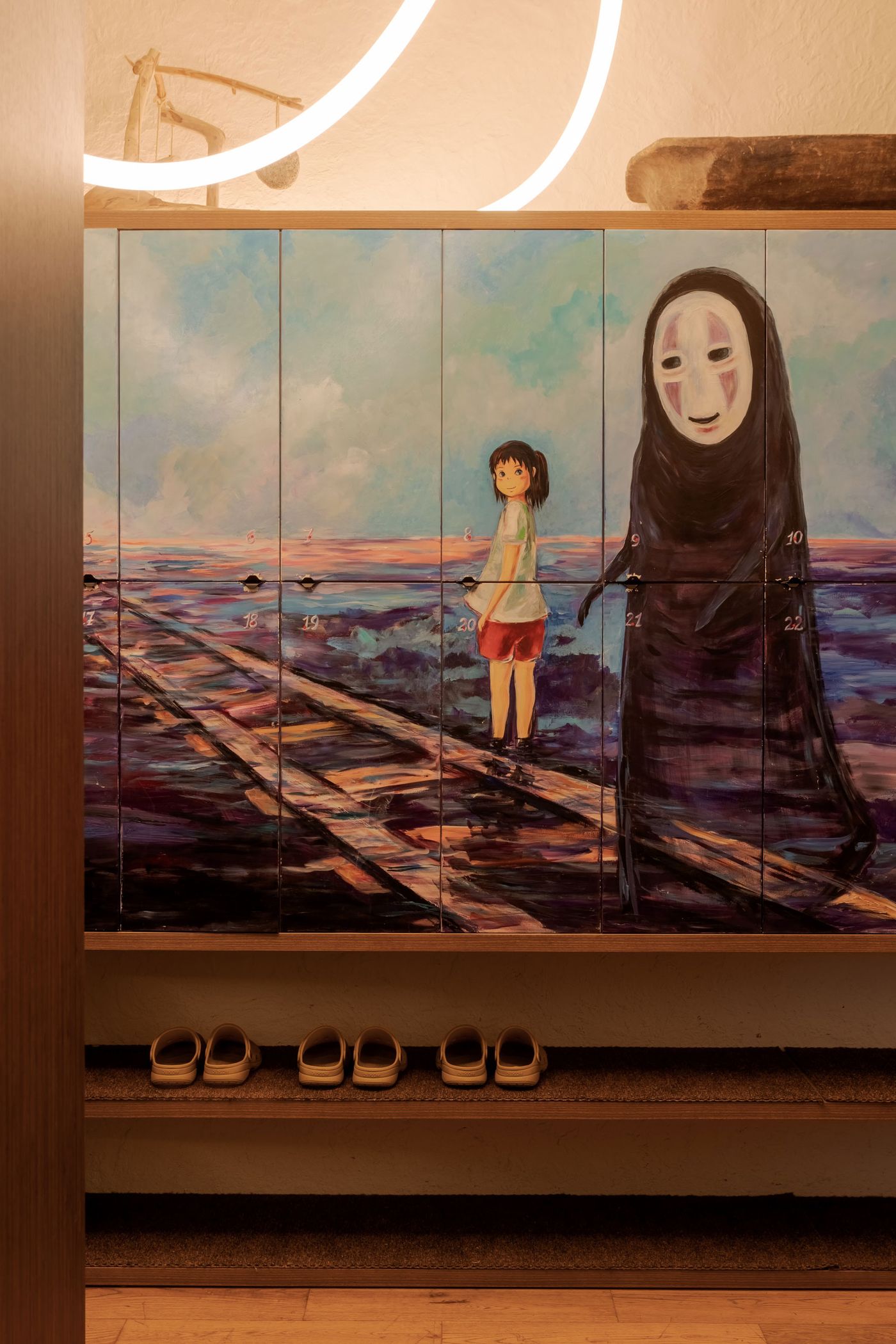 Colorful lockers adorned with a mural depicting characters from Hayao Miyazaki's animated film Spirited Away on a railway, contrasting with a dark figure in a flowing garment beside her. Colorful lockers adorned with a mural depicting characters from Hayao Miyazaki's animated film Spirited Away on a railway, contrasting with a dark figure in a flowing garment beside her.