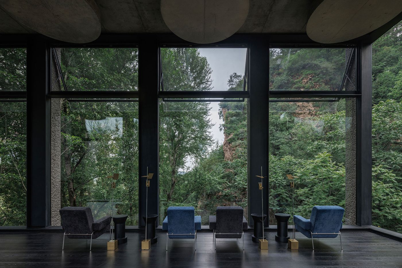 Modern lounge with blue and gray chairs, framed by large windows overlooking lush greenery and rocky cliffs outside. Modern lounge with blue and gray chairs, framed by large windows overlooking lush greenery and rocky cliffs outside.