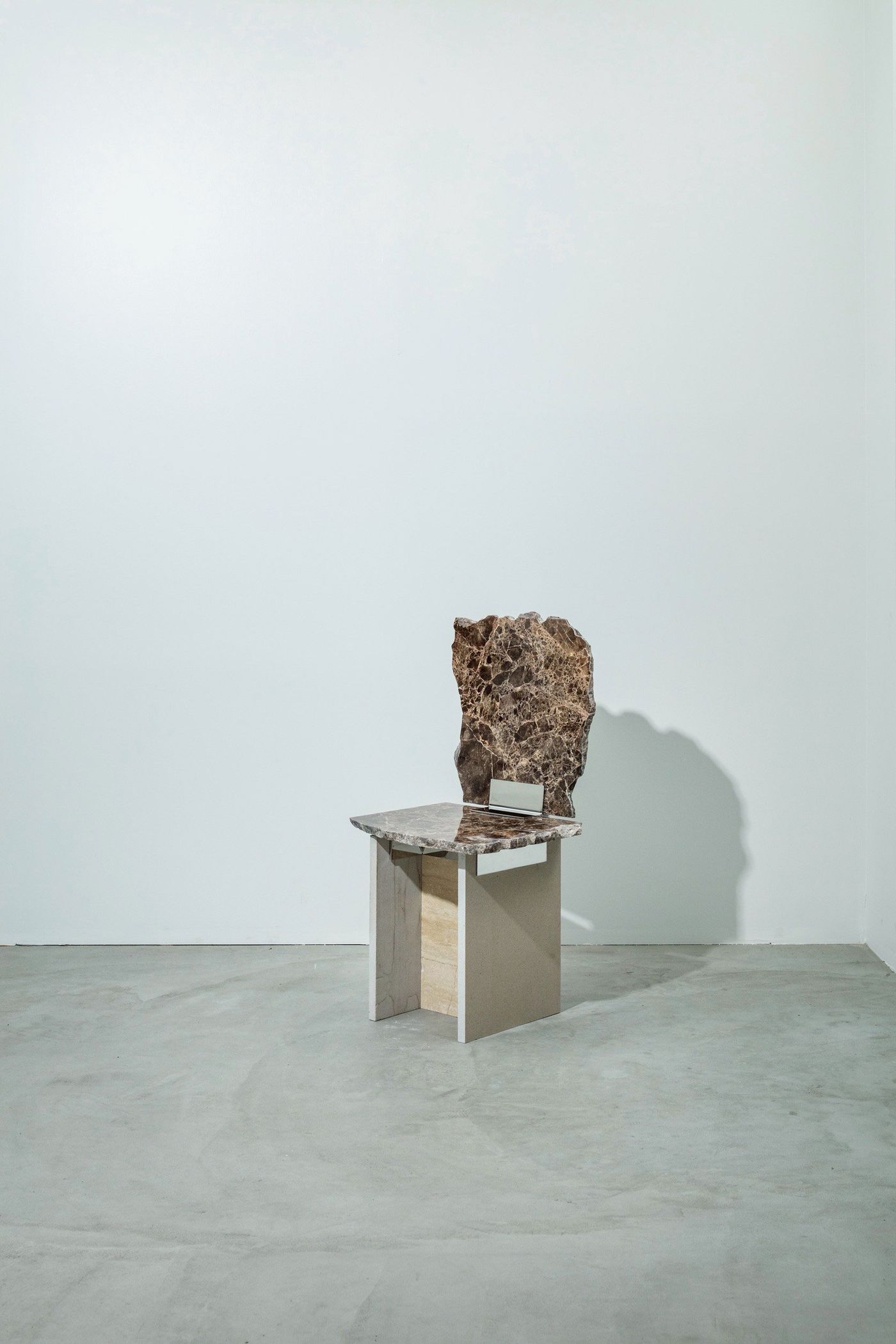 A minimalist chair with a large, irregular stone backrest sits on a simple, modern base in a bright, empty room.