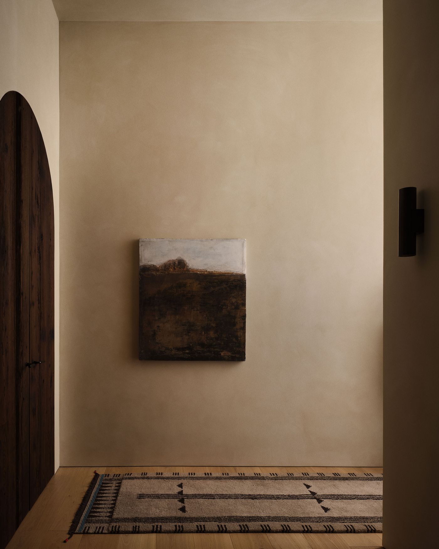 A serene hallway features a muted landscape painting, rustic wooden door, and a patterned rug on light wood flooring.