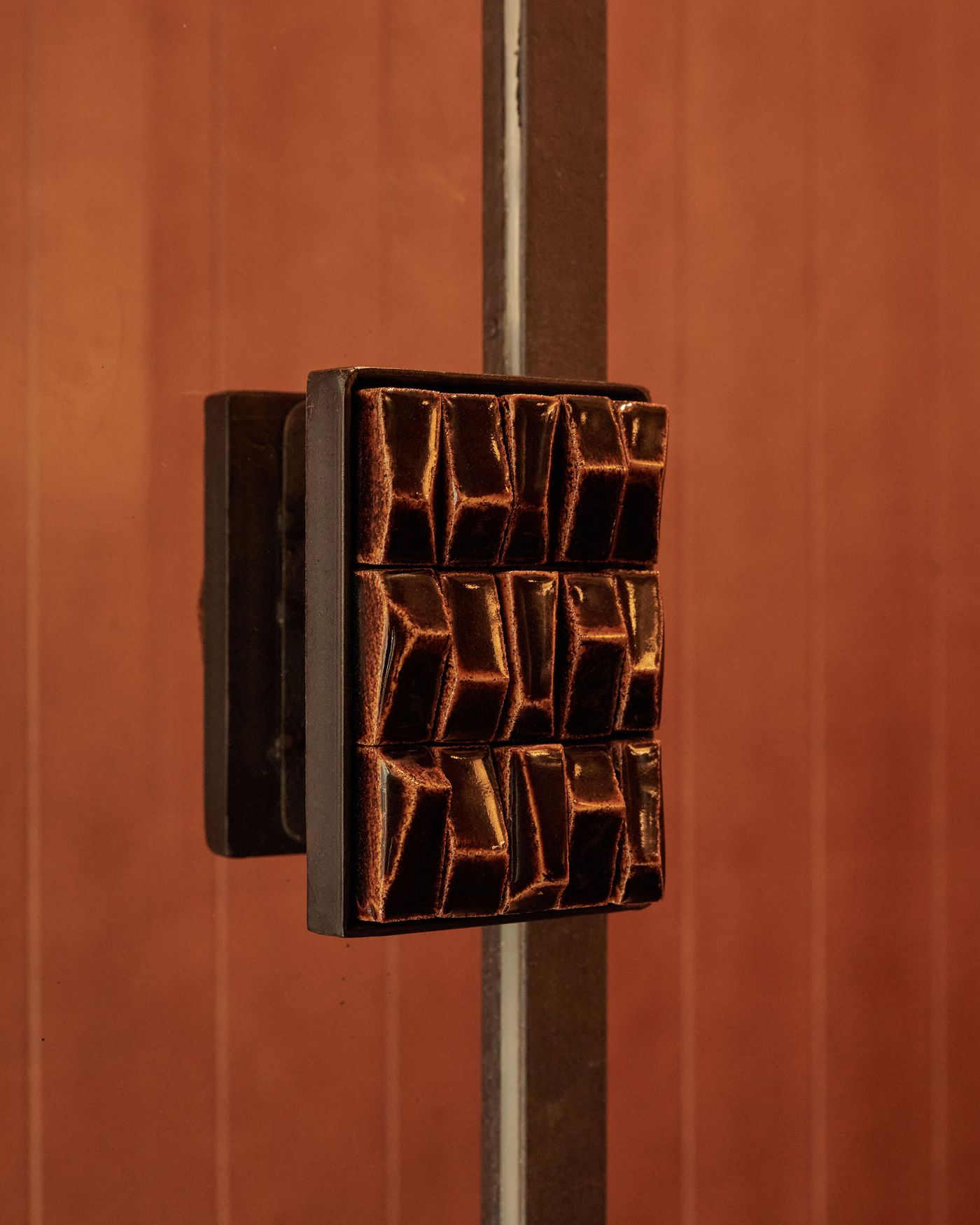 A close-up of a striking, vintage-inspired door handle crafted from dark, glazed ceramic. Its geometric, grid-like relief contrasts with the smooth, warm terracotta-toned door and a sleek metal frame, creating a tactile and visually compelling entrance detail.