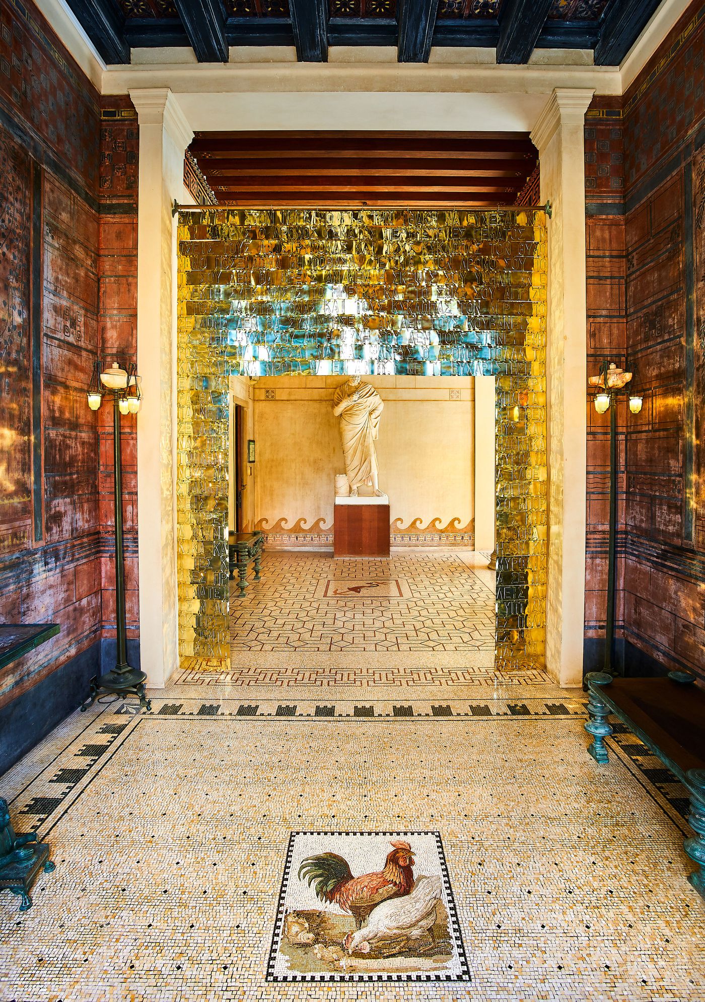 A Grecian entrance hall at Villa Kerylos, featuring a floor mosaic of a rooster and hen. A modern art installation by Gabriel Leger, made of hanging golden metal sheets, frames the doorway. The space is richly decorated with murals of waves and patterns, illuminated by a warm, ethereal glow. A Grecian entrance hall at Villa Kerylos, featuring a floor mosaic of a rooster and hen. A modern art installation by Gabriel Leger, made of hanging golden metal sheets, frames the doorway. The space is richly decorated with murals of waves and patterns, illuminated by a warm, ethereal glow.