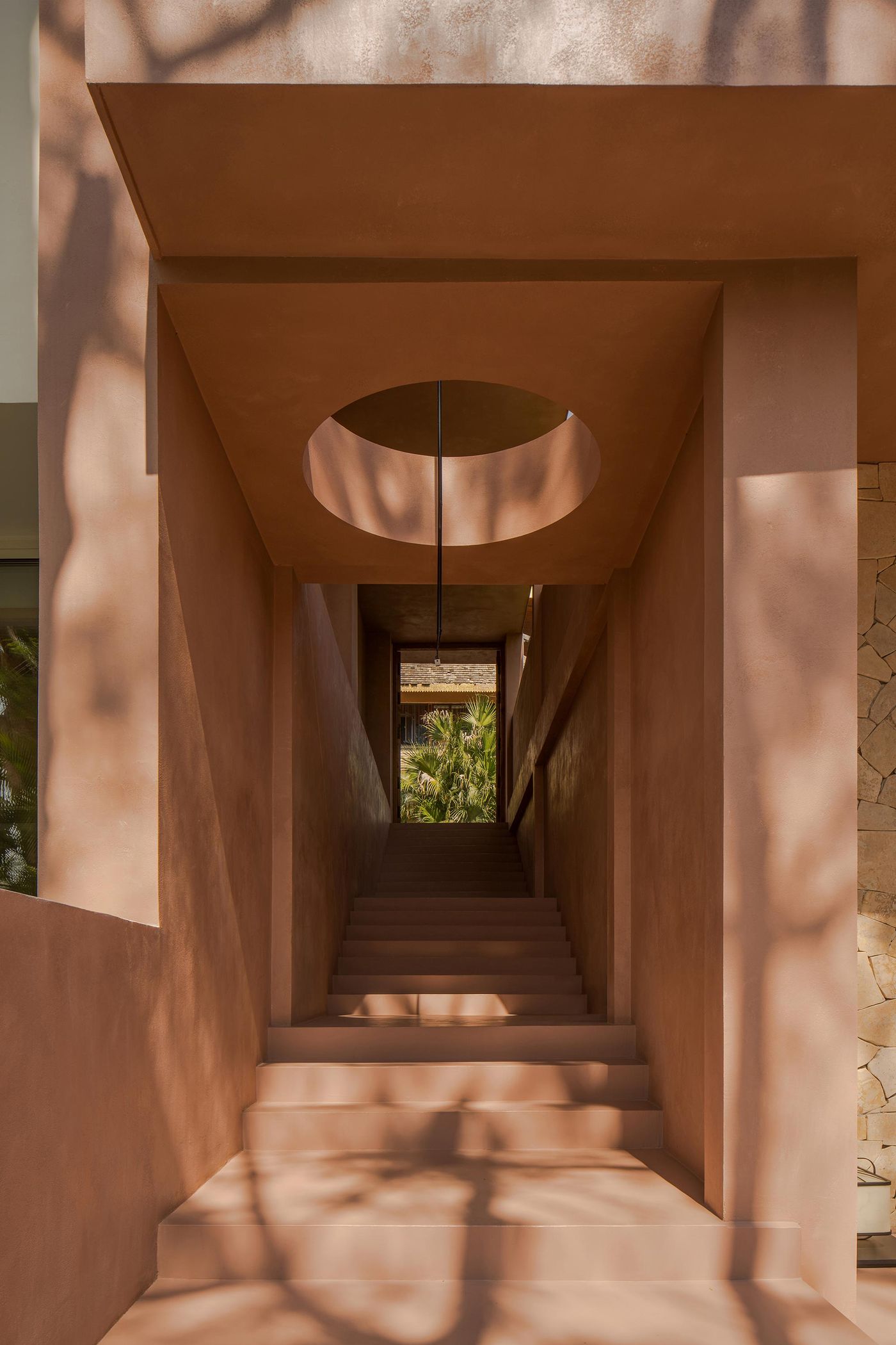 A warm-toned corridor with steps leading up, featuring a round opening overhead and lush greenery visible at the end. A warm-toned corridor with steps leading up, featuring a round opening overhead and lush greenery visible at the end.