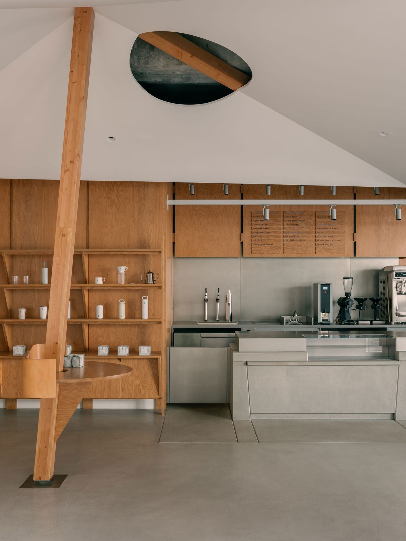 A minimalist café counter in Blue Bottle Coffee Shanghai blends cool grey concrete with warm oak cabinetry and a sculptural timber column, set beneath a sharply faceted ceiling pierced by a circular skylight.