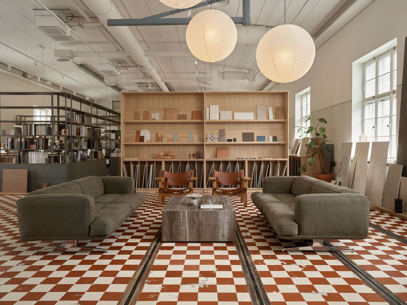 A spacious industrial-chic office lounge features two olive-green sofas and a marble coffee table on a distinctive red and white checkerboard floor. Large paper lanterns illuminate the space, complementing the minimalist wooden shelving showcasing design samples. A spacious industrial-chic office lounge features two olive-green sofas and a marble coffee table on a distinctive red and white checkerboard floor. Large paper lanterns illuminate the space, complementing the minimalist wooden shelving showcasing design samples.