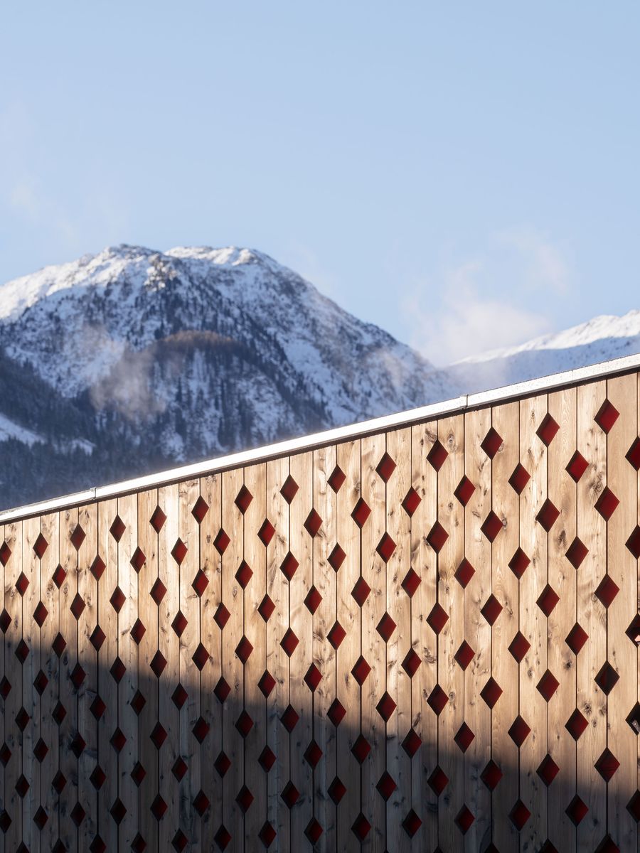 A striking perforated timber facade creates an intricate diamond pattern against snow-capped alpine peaks. The rhythmic geometric cutouts in warm wood contrast beautifully with the pristine mountain landscape, exemplifying contemporary Alpine vernacular architecture that harmonizes built environment with natural grandeur. A striking perforated timber facade creates an intricate diamond pattern against snow-capped alpine peaks. The rhythmic geometric cutouts in warm wood contrast beautifully with the pristine mountain landscape, exemplifying contemporary Alpine vernacular architecture that harmonizes built environment with natural grandeur.