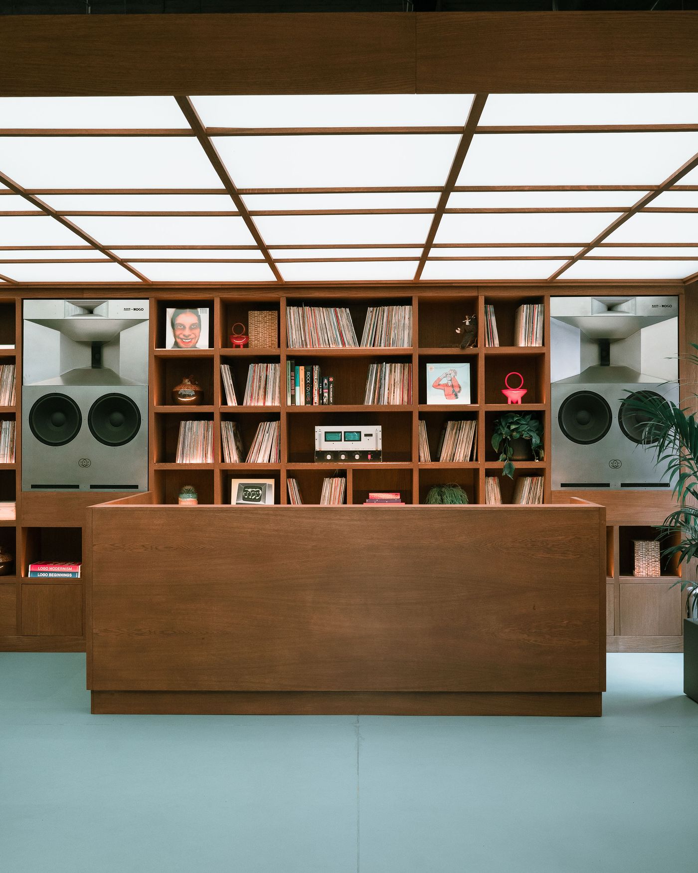 A wooden reception desk in front of shelves filled with vinyl records and large speakers, against a softly illuminated ceiling. A wooden reception desk in front of shelves filled with vinyl records and large speakers, against a softly illuminated ceiling.