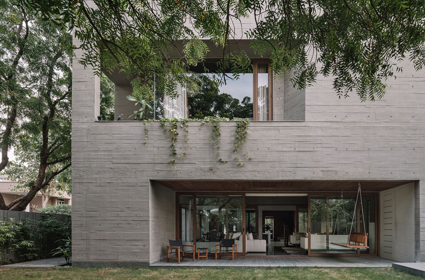 The facade of MS House showcases a harmonious blend of brutalist architecture and biophilic design. Raw, textured concrete walls are softened by cascading green vines and the lush canopy of surrounding trees. A ground-level outdoor living space with comfortable chairs and a wooden swing is visible through large glass doors, seamlessly connecting the interior with nature.