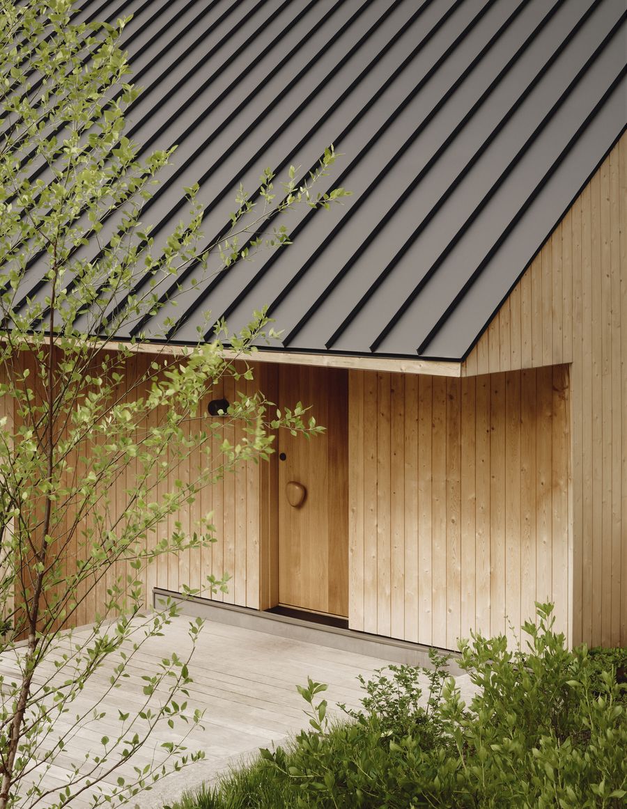A modern wooden house features a sleek black metal roof, surrounded by greenery and showcasing a unique rock-shaped wooden door handle. A modern wooden house features a sleek black metal roof, surrounded by greenery and showcasing a unique rock-shaped wooden door handle.