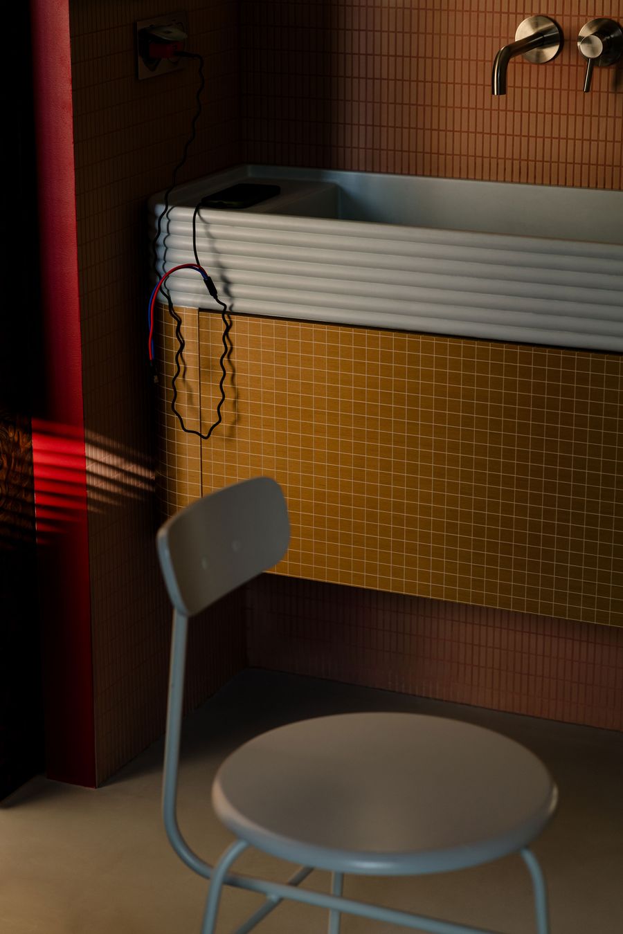 A moody bathroom vignette contrasts earthy terracotta and mustard grid tiles with a ribbed white sink basin. A sculptural steel faucet and looping electrical cords accentuate the utilitarian, postmodern edge of the composition, softened by warm, directional light filtering through blinds. A moody bathroom vignette contrasts earthy terracotta and mustard grid tiles with a ribbed white sink basin. A sculptural steel faucet and looping electrical cords accentuate the utilitarian, postmodern edge of the composition, softened by warm, directional light filtering through blinds.
