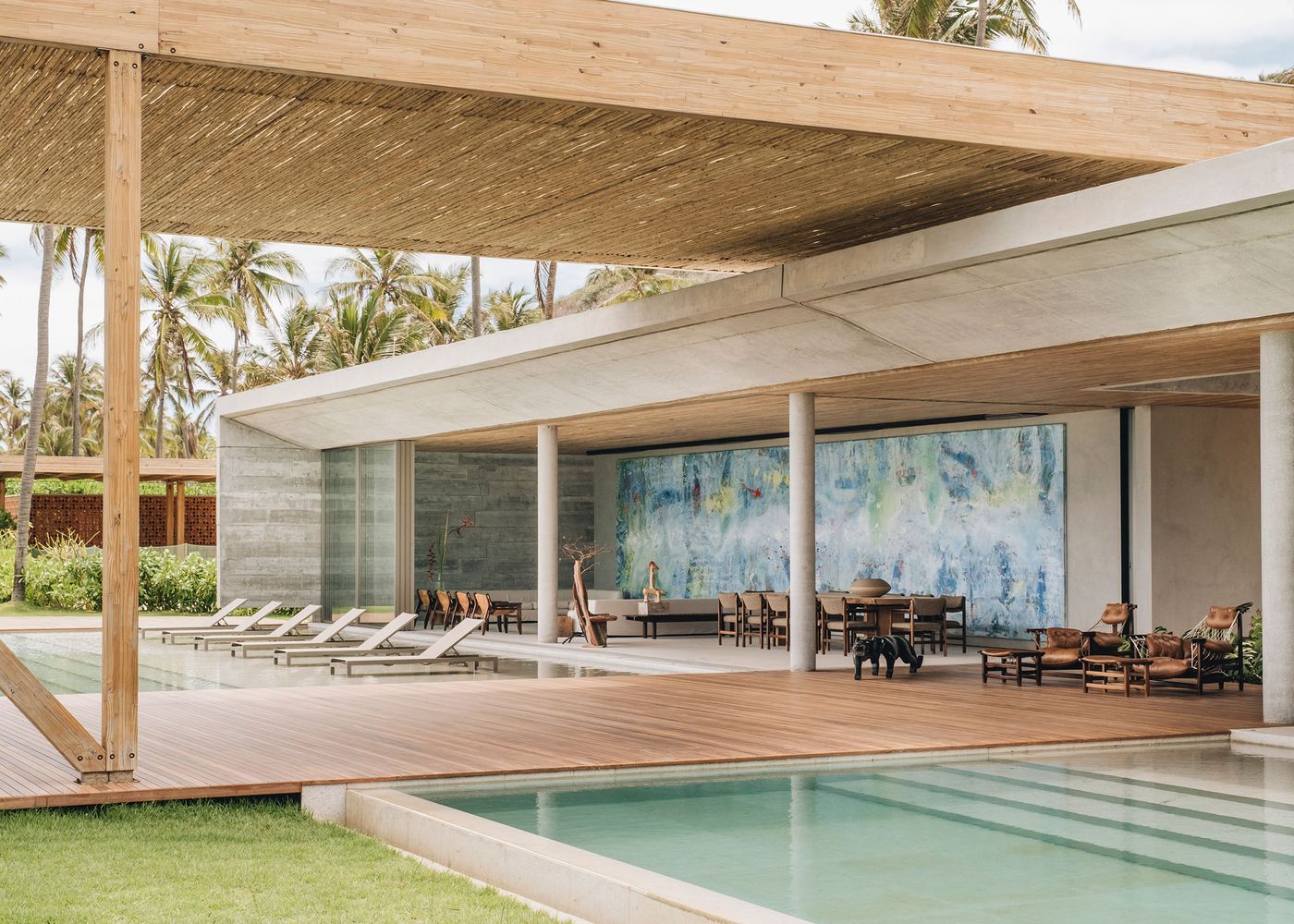 Modern villa with a wooden overhang, poolside seating, and an abstract blue mural, surrounded by palm trees and lush greenery.