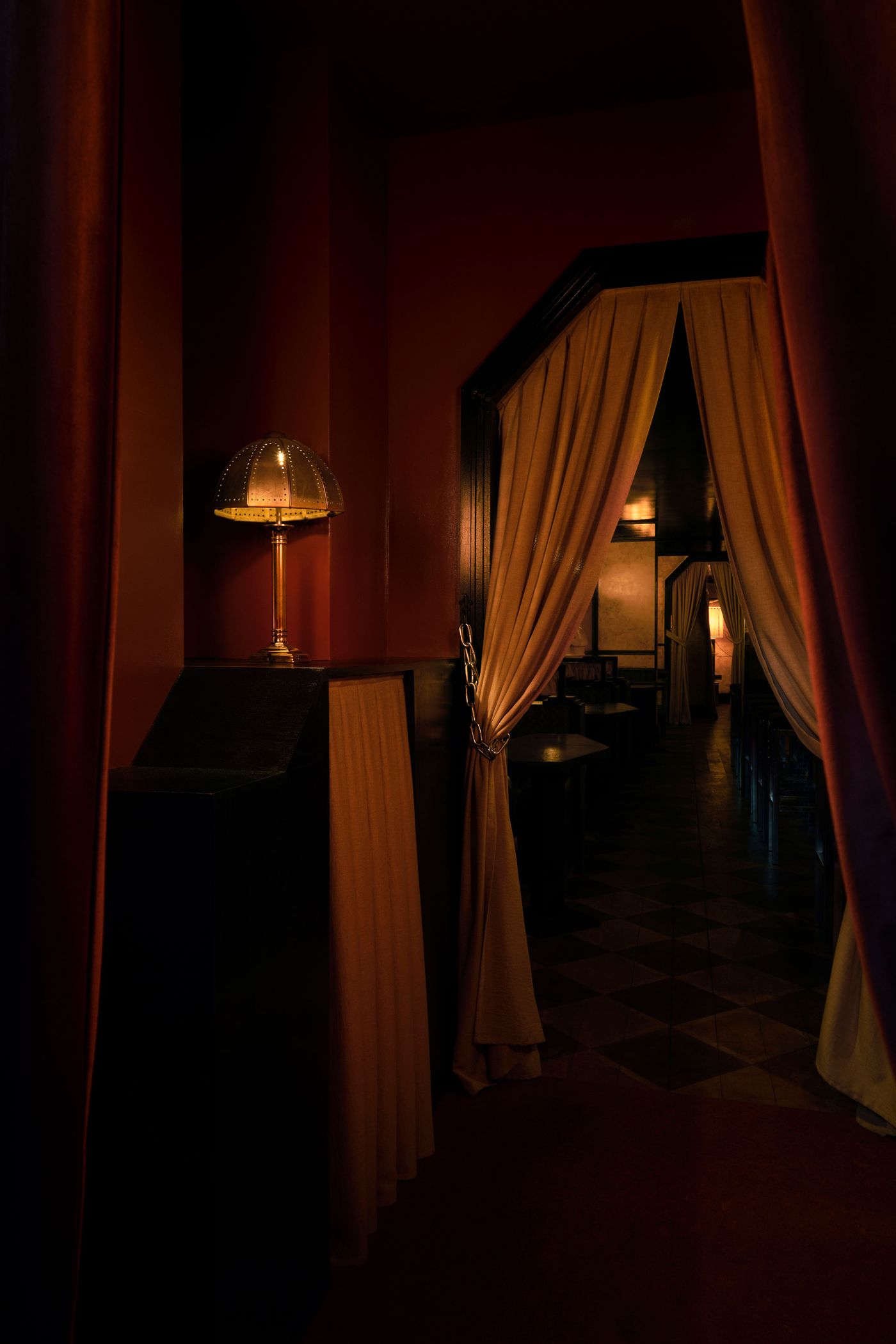 A richly atmospheric, vintage-retro interior at The Roses of Elagabalus in London, bathed in deep red and amber tones. Heavy drapes in a warm, muted orange frame an arched doorway, hinting at hidden spaces. A petite, ornate metallic table lamp casts a warm glow, enhancing the intimate, secretive ambiance of this opulent space. A richly atmospheric, vintage-retro interior at The Roses of Elagabalus in London, bathed in deep red and amber tones. Heavy drapes in a warm, muted orange frame an arched doorway, hinting at hidden spaces. A petite, ornate metallic table lamp casts a warm glow, enhancing the intimate, secretive ambiance of this opulent space.