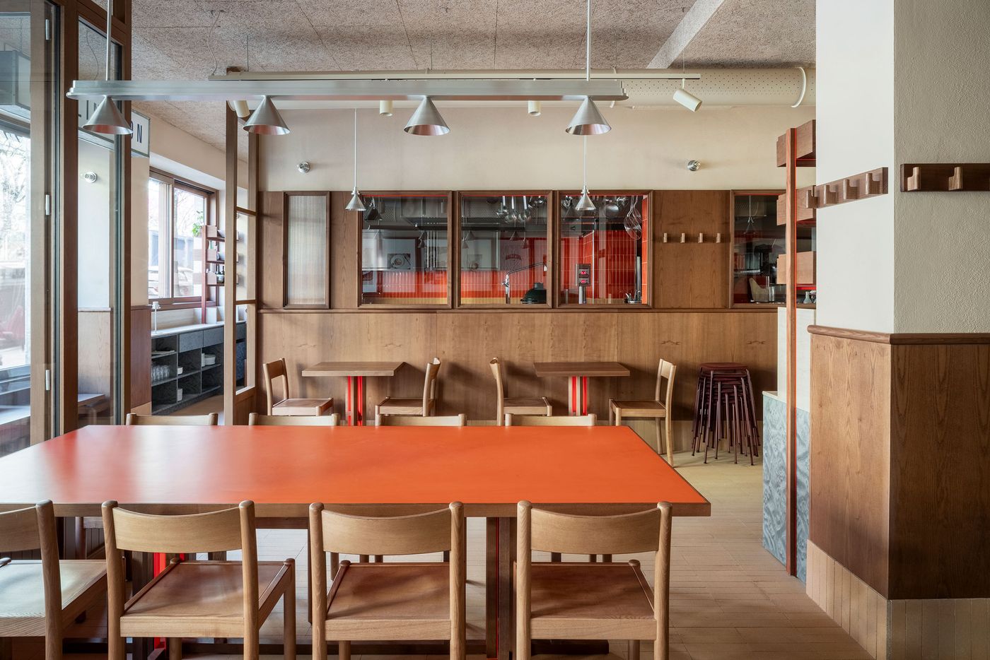 The main dining area of Santo Palato by Naessi Studio, defined by a large, bold orange communal table with subtle red legs, contrasting with the warm wooden chairs. The industrial-chic aesthetic features exposed ceiling pipes and minimalist pendant lights. Behind the table, smaller wooden tables with red bases are visible, contributing to a modern, convivial atmosphere. The main dining area of Santo Palato by Naessi Studio, defined by a large, bold orange communal table with subtle red legs, contrasting with the warm wooden chairs. The industrial-chic aesthetic features exposed ceiling pipes and minimalist pendant lights. Behind the table, smaller wooden tables with red bases are visible, contributing to a modern, convivial atmosphere.