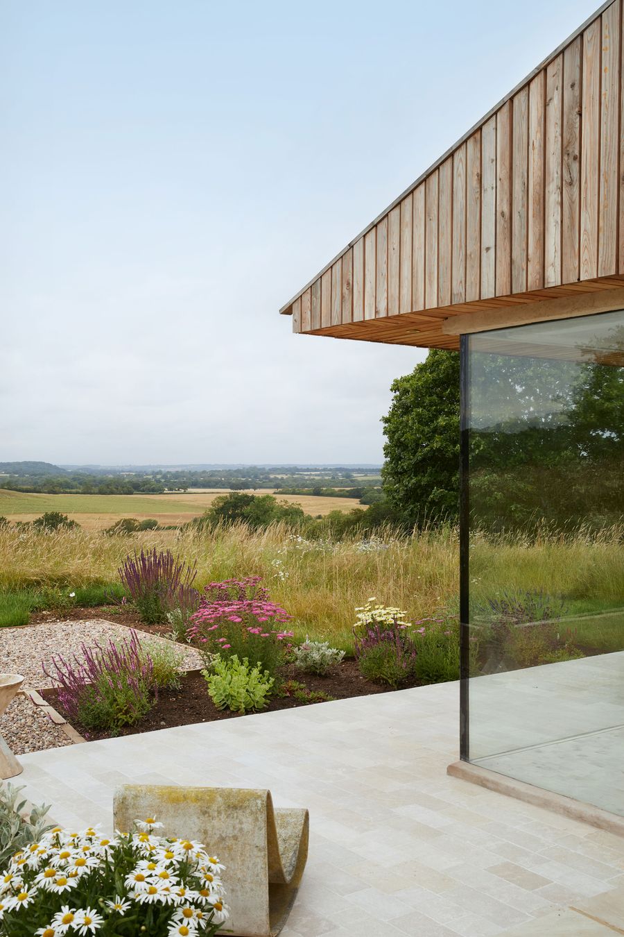This biophilic design features a modern structure by Hutch Design blending into a pastoral landscape. A minimalist stone patio with a unique curved bench and planted garden beds foregrounds a wall of glass, reflecting the expansive golden fields and distant trees under an overcast sky, evoking a serene rural retreat. This biophilic design features a modern structure by Hutch Design blending into a pastoral landscape. A minimalist stone patio with a unique curved bench and planted garden beds foregrounds a wall of glass, reflecting the expansive golden fields and distant trees under an overcast sky, evoking a serene rural retreat.
