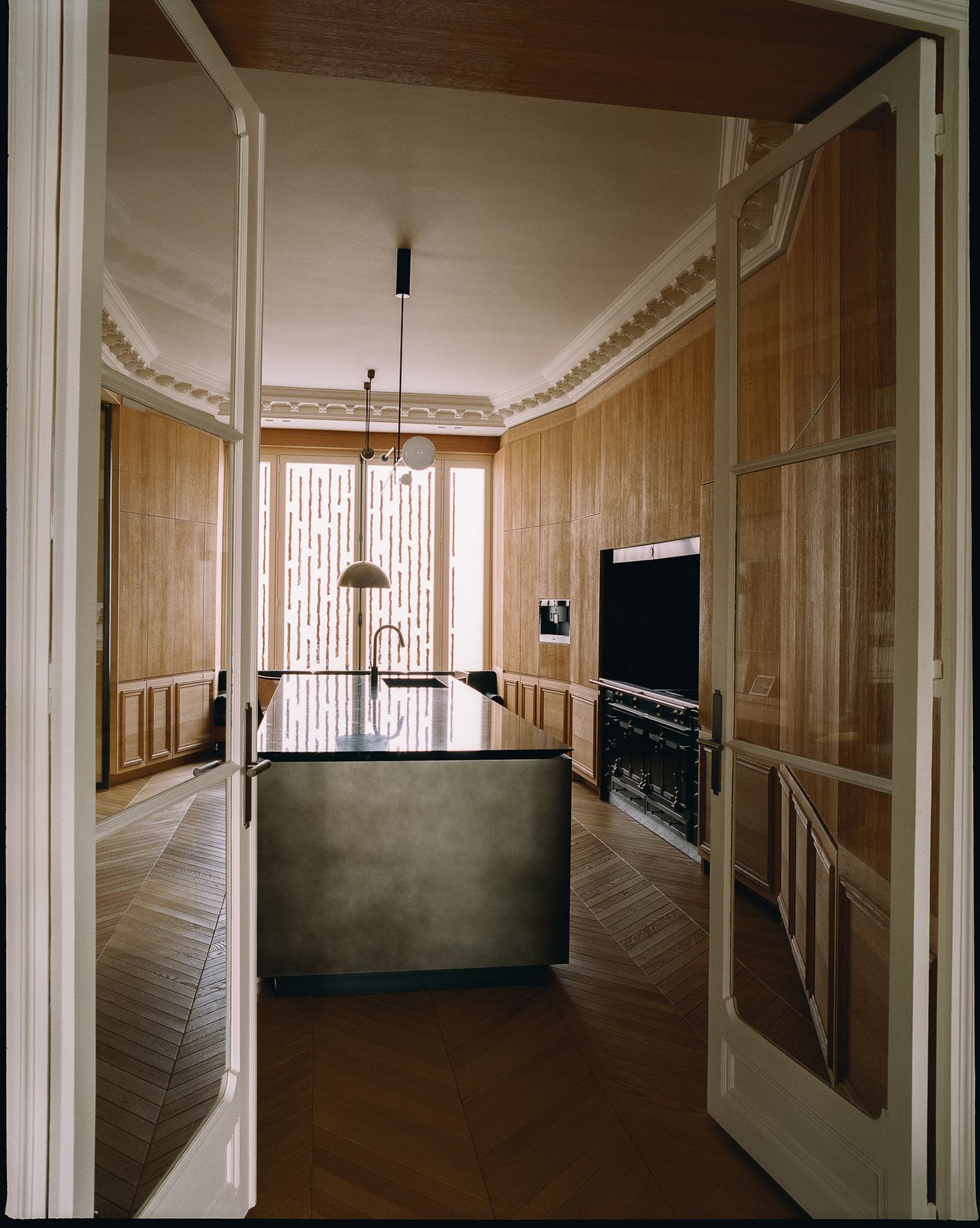 A sophisticated Parisian kitchen, designed by Isabelle Stanislas, features custom oak paneling and a large, dark-hued central island with a sleek countertop. The herringbone wooden floor adds classic warmth, contrasting with the minimalist black cabinetry and the industrial-chic pendant light, viewed through elegant French doors.