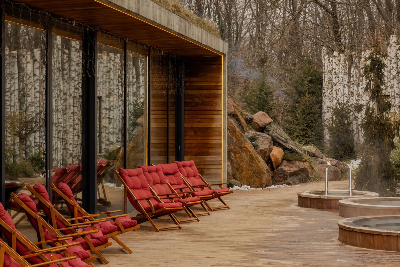 A serene wood deck featuring red cushioned lounge chairs, framed by large windows and a backdrop of birch trees and rocky landscape. A serene wood deck featuring red cushioned lounge chairs, framed by large windows and a backdrop of birch trees and rocky landscape.