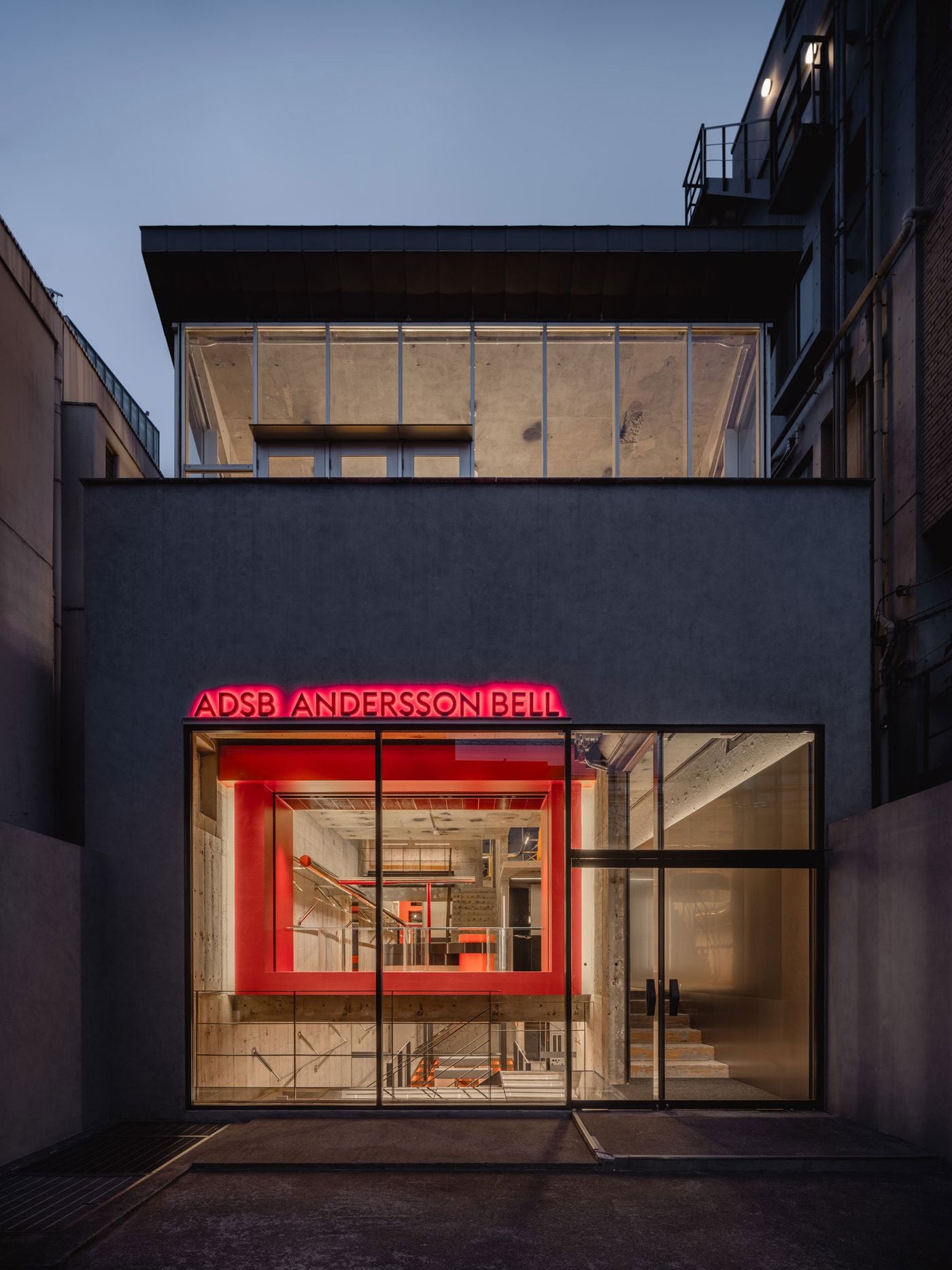 At dusk, ANDERSSON BELL Tokyo presents a restrained concrete façade punctuated by a glowing red sign and a large glazed opening. The vivid red portal of the ground floor is clearly visible from outside, while the glazed upper level reveals the preserved concrete structure above, turning the building into a layered urban display.