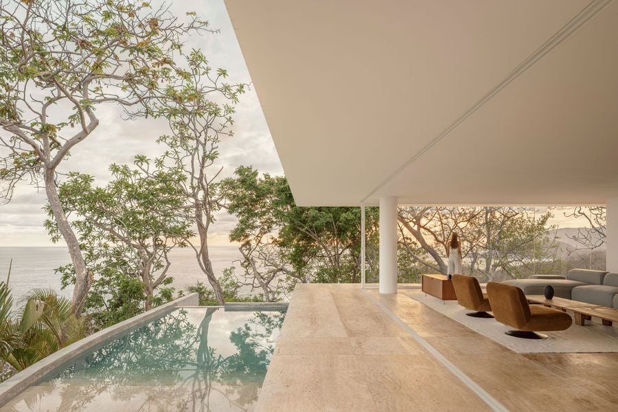 A thin white roof slab cuts a sharp diagonal against the sky, sheltering the living area beside a still pool that mirrors the surrounding trees. Pale paving runs uninterrupted across the flush sliding-door track, while a woman gazes through the glazing towards wooded hills in soft, low light.