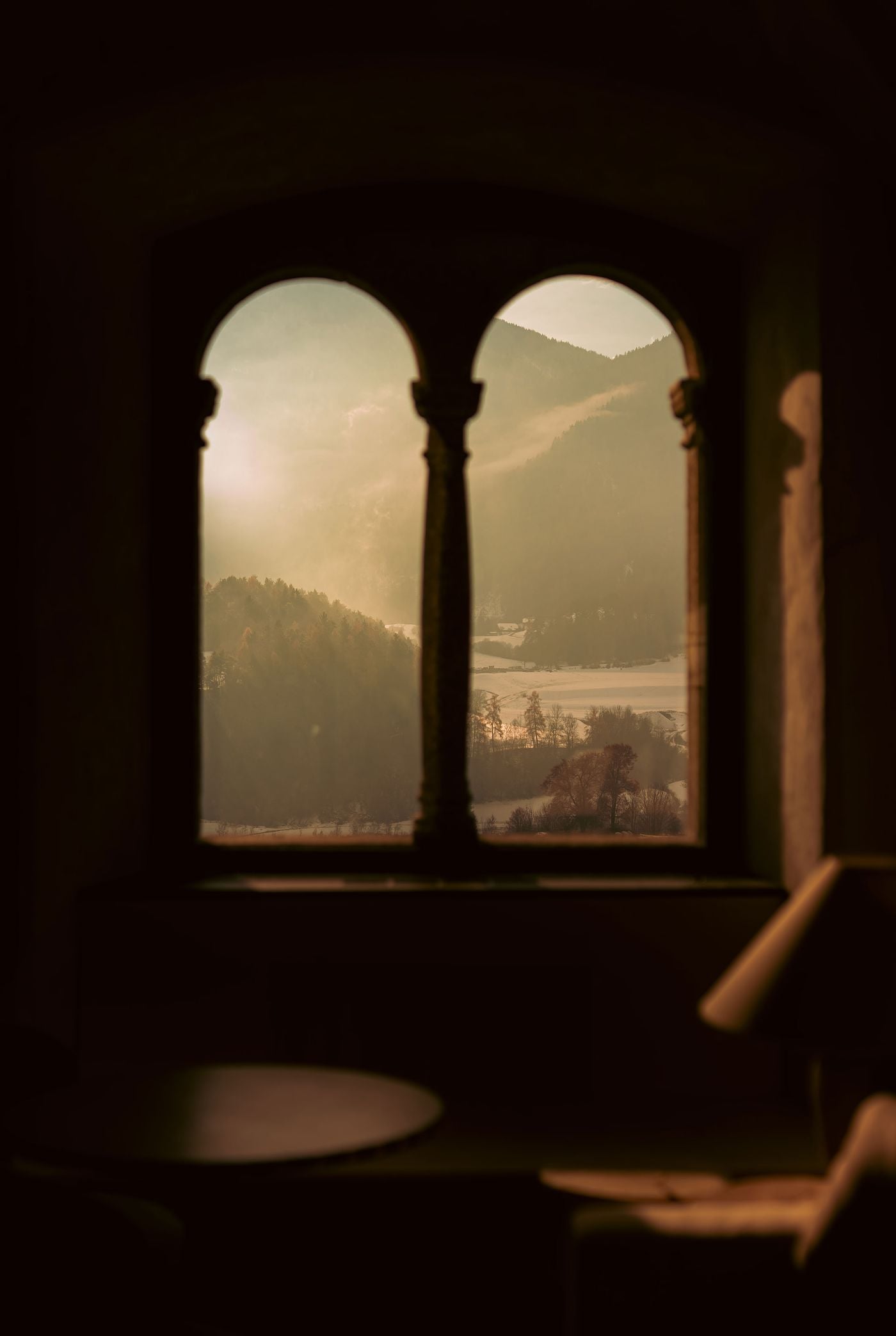 A twin-arched Romanesque window, divided by a slender column with carved capitals, frames a misty valley view at Castel Badia. The deep stone reveal and darkened interior, with a table and lamp in soft silhouette, heighten the contrast with the hazy light outside. A twin-arched Romanesque window, divided by a slender column with carved capitals, frames a misty valley view at Castel Badia. The deep stone reveal and darkened interior, with a table and lamp in soft silhouette, heighten the contrast with the hazy light outside.