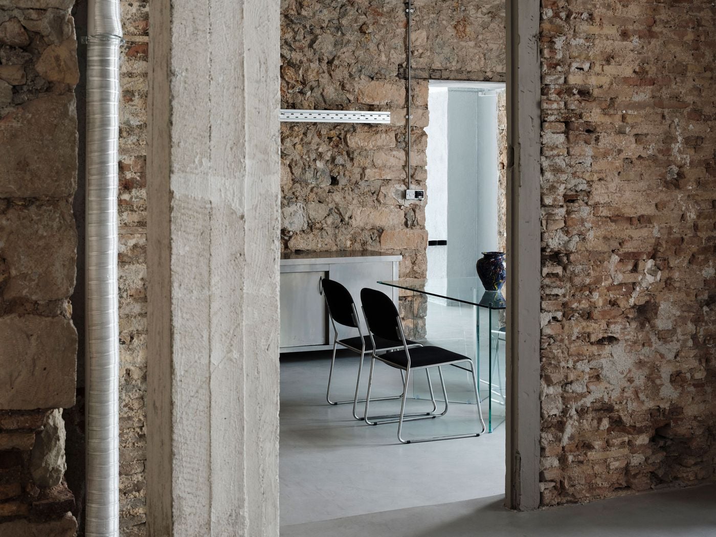 Layers of stone, brick and concrete frame a meeting area furnished with slender black chairs and a transparent glass table. Exposed ductwork and electrical conduits reinforce the building’s industrial character, while the restrained contemporary furniture allows the richly textured fabric of the former port warehouse to remain visually dominant. Layers of stone, brick and concrete frame a meeting area furnished with slender black chairs and a transparent glass table. Exposed ductwork and electrical conduits reinforce the building’s industrial character, while the restrained contemporary furniture allows the richly textured fabric of the former port warehouse to remain visually dominant.