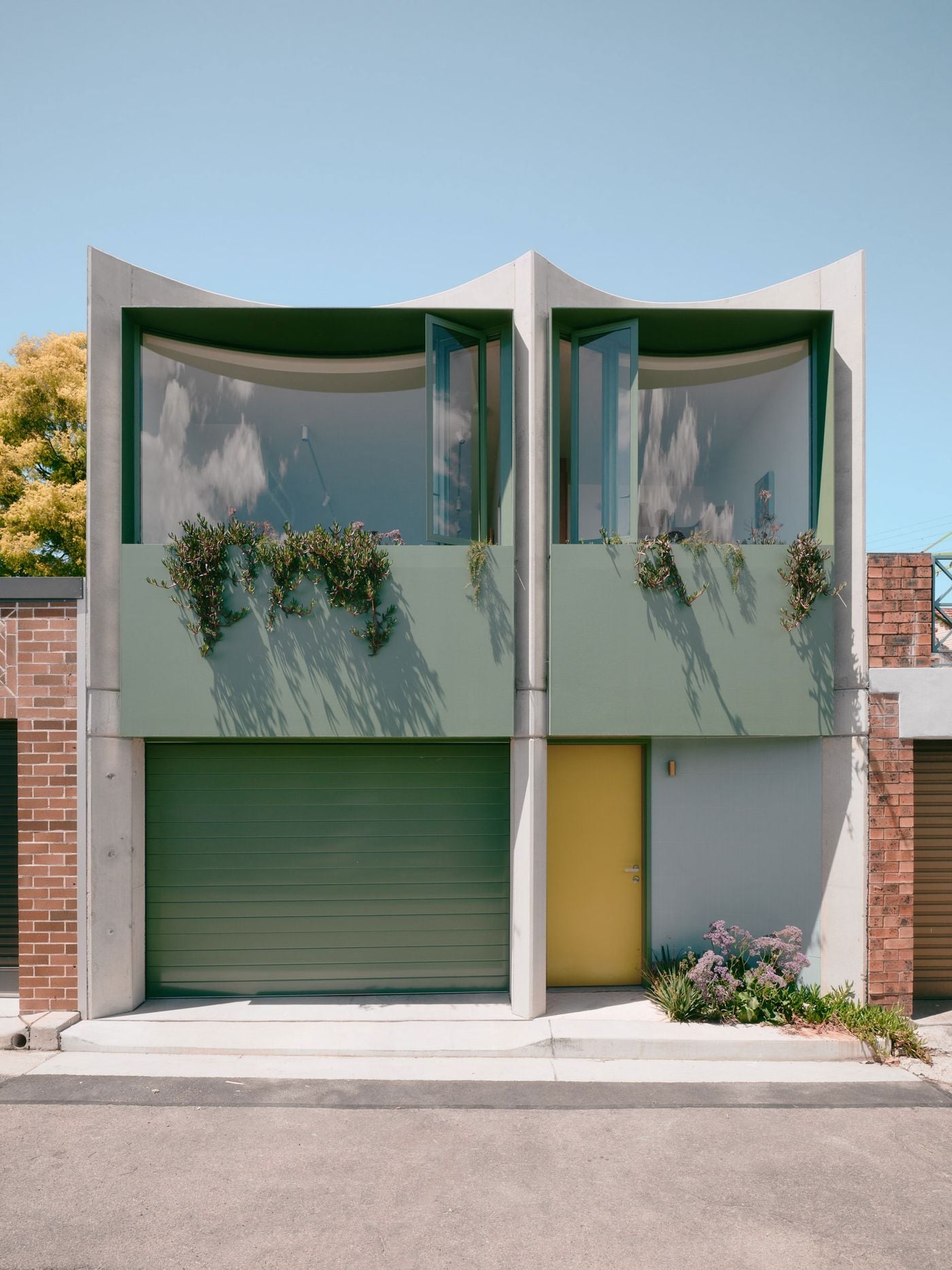A frontal view of Denj Lane highlights Carter Williamson Architects’ expressive laneway elevation, where twin concave concrete bays frame sage-green glazing and planter boxes. Matching garage doors unify the composition, while a mustard-yellow entrance and small patch of flowering landscape introduce a distinctly domestic note to the otherwise utilitarian streetscape. A frontal view of Denj Lane highlights Carter Williamson Architects’ expressive laneway elevation, where twin concave concrete bays frame sage-green glazing and planter boxes. Matching garage doors unify the composition, while a mustard-yellow entrance and small patch of flowering landscape introduce a distinctly domestic note to the otherwise utilitarian streetscape.